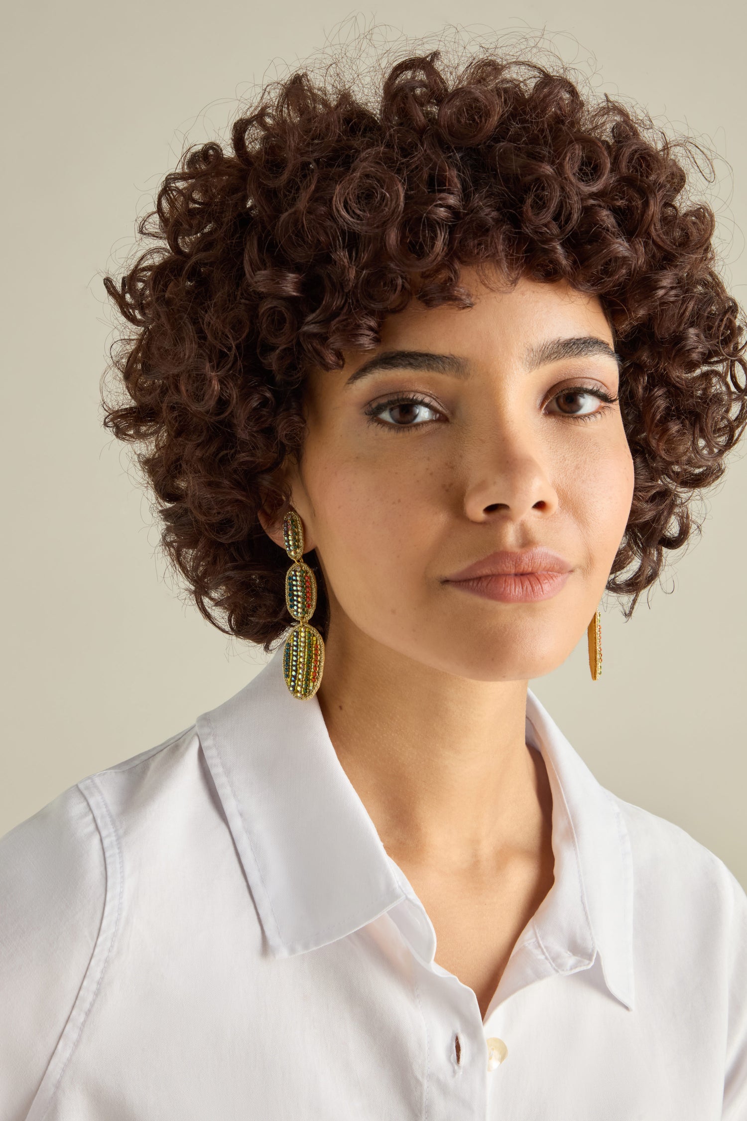 A curly-haired person in a white shirt gazes at the camera, highlighting Hand Embroidered Crystal Bead Earrings with intricate multi-colored beadwork and a stunning drop design.