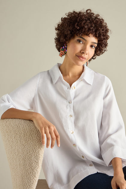 A person with curly hair and Hand Beaded Frida Earrings sits on a chair, wearing a white button-up shirt.