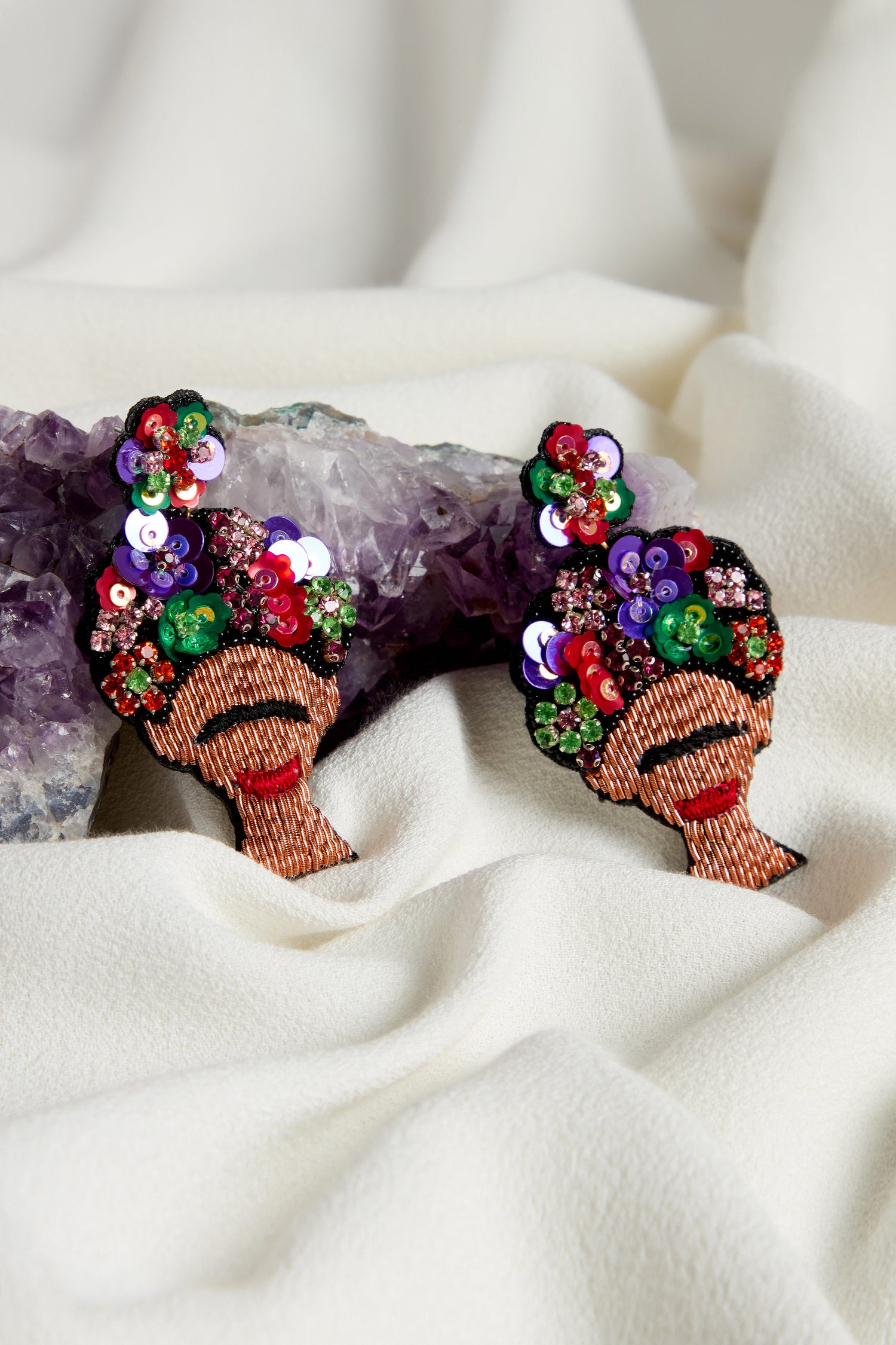 Hand Beaded Frida Earrings with face designs and bright floral headpieces are displayed on white fabric next to an amethyst.