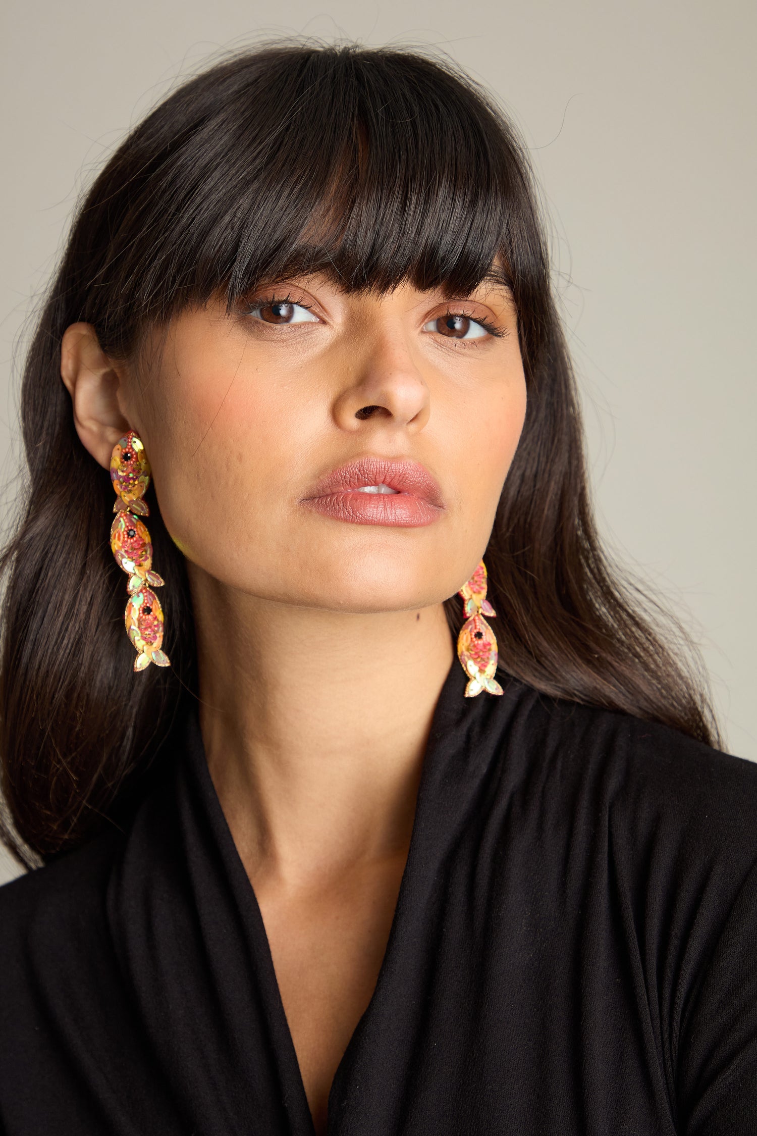 A woman with long dark hair and bangs wears the Hand Beaded Poisson Earrings, featuring ornate gold and pink designs adorned with iridescent beads and sequins, as she looks directly at the camera. She is dressed in a black outfit against a plain background.