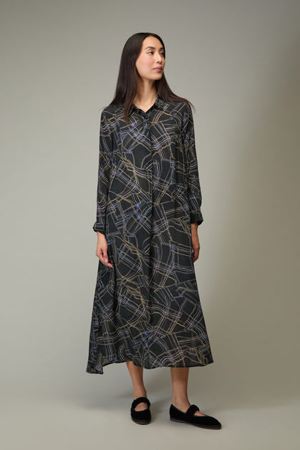 A woman stands slightly sideways, wearing the Wavy Lines Shirt Dress—an ankle-length, long-sleeved dress with an abstract print—and black shoes against a plain background.