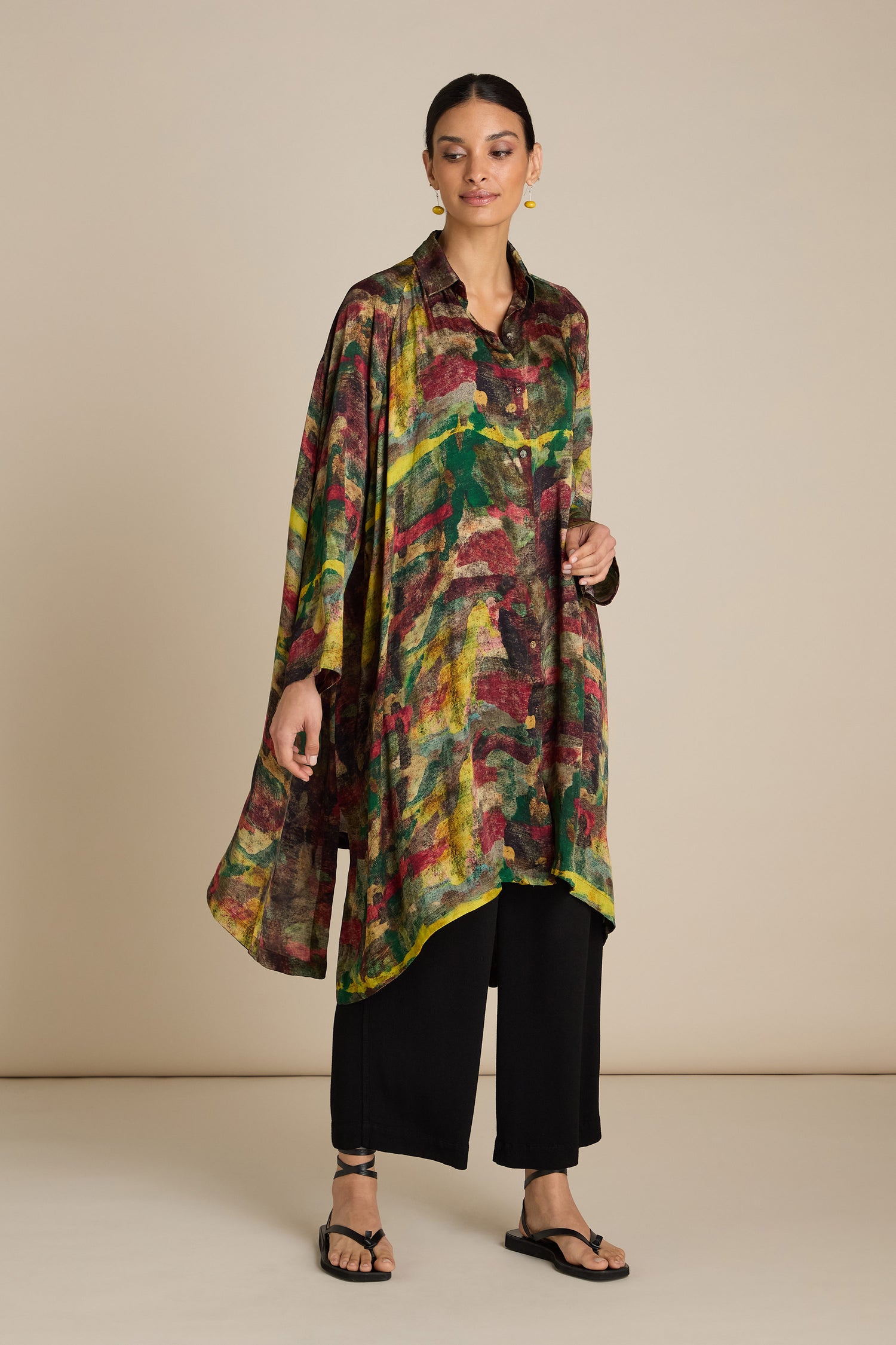 A woman stands against a plain background wearing a colorful, loose-fitting Abstract Camo Shirt Dress with abstract patterns, black pants, and sandals. Her painterly inspired print dress adds an artistic flair as she looks slightly to the side with her hair pulled back.