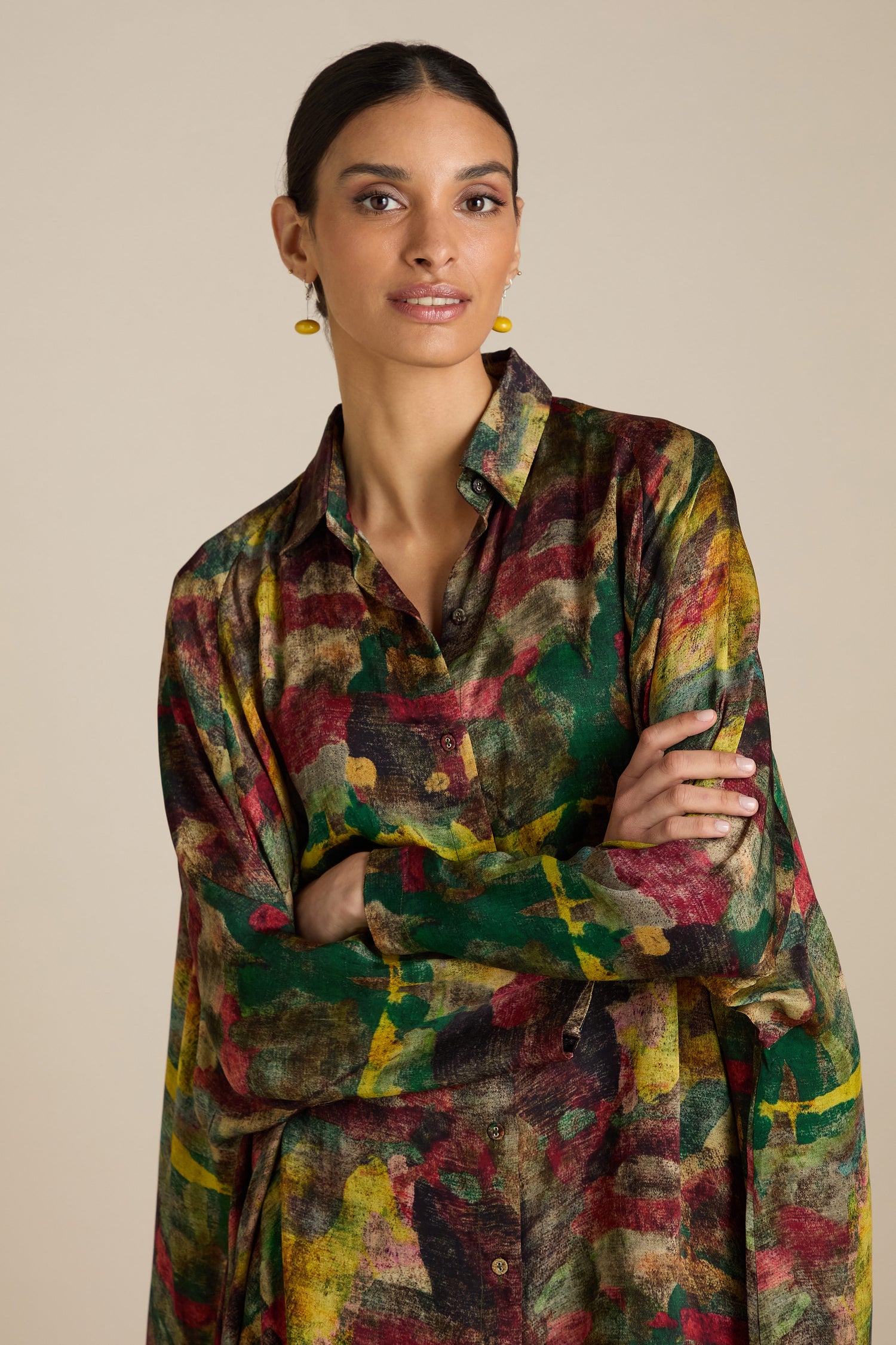 A person with dark hair is wearing an Abstract Camo Shirt Dress and yellow earrings, standing with arms crossed against a plain beige background.