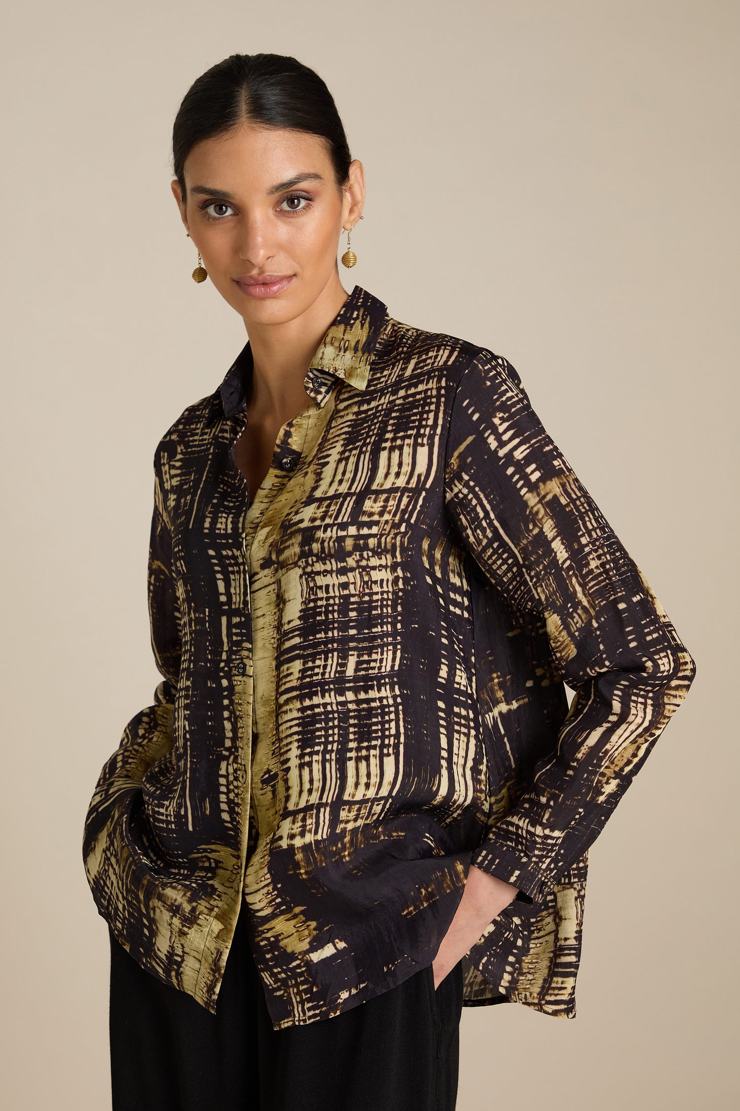 A person wearing a long-sleeved, Cross Hatch Shirt with earthy tones and black pants stands against a plain background. The relaxed fit of their outfit, featuring silky Viscose fabric, complements their dark, pulled-back hair as they look directly at the camera, perfecting a daytime look.