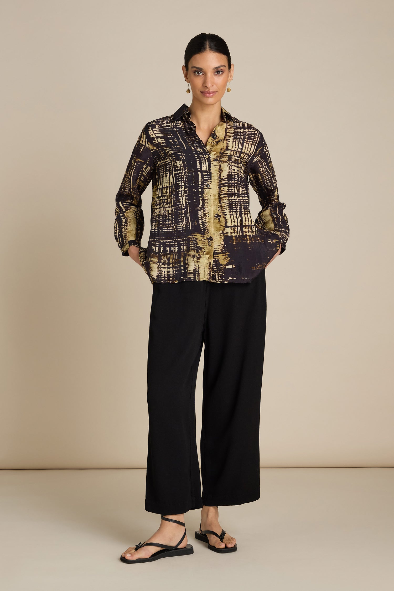 A woman stands against a beige background wearing a Cross Hatch Shirt, black wide-legged pants with a relaxed fit, and sandals, perfect for a stylish daytime look.