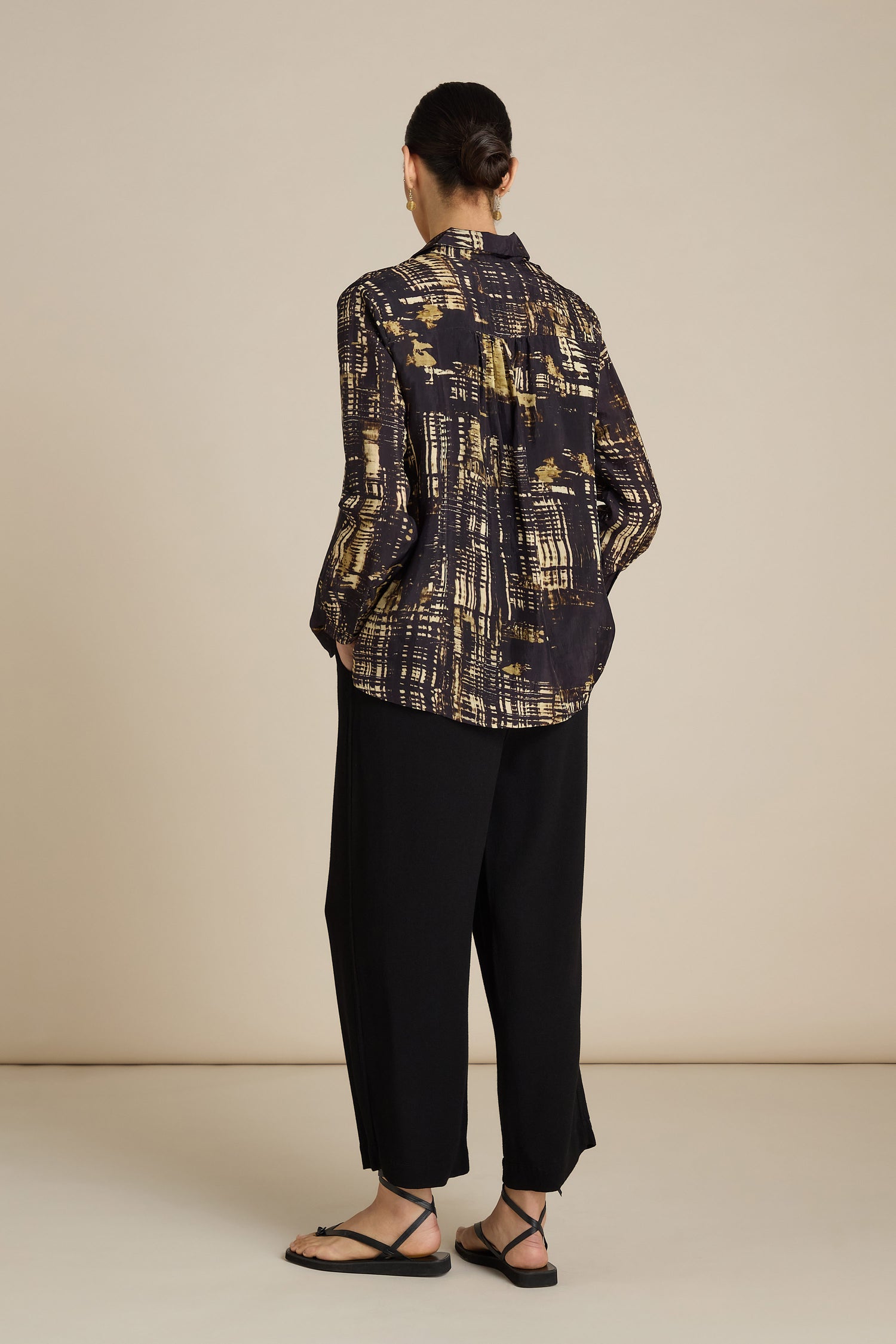 Person stands facing away, wearing a long-sleeve Cross Hatch Shirt made of silky Viscose, black loose-fit pants, and black sandals. Hair is tied in a bun. Neutral background emphasizes this relaxed fit and ideal daytime look.