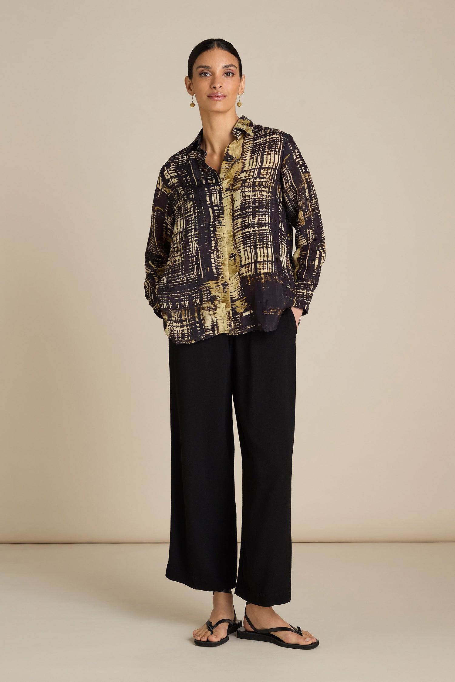 A person stands against a neutral background, wearing a black and gold patterned Cross Hatch Shirt, black wide-leg pants for a relaxed fit, and black sandals, perfect for a chic daytime look.