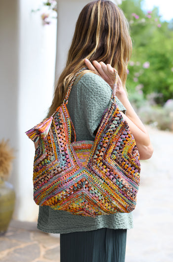 A person in a green outfit graces the scene, holding a vibrant Handwoven Bohemian Raffia Bag adorned with a zigzag pattern.