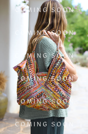 Dressed in a textured green outfit, someone holds a Handwoven Bohemian Raffia Bag. Surrounded by lush greenery and elegant architecture, the text softly announces, "COMING SOON.