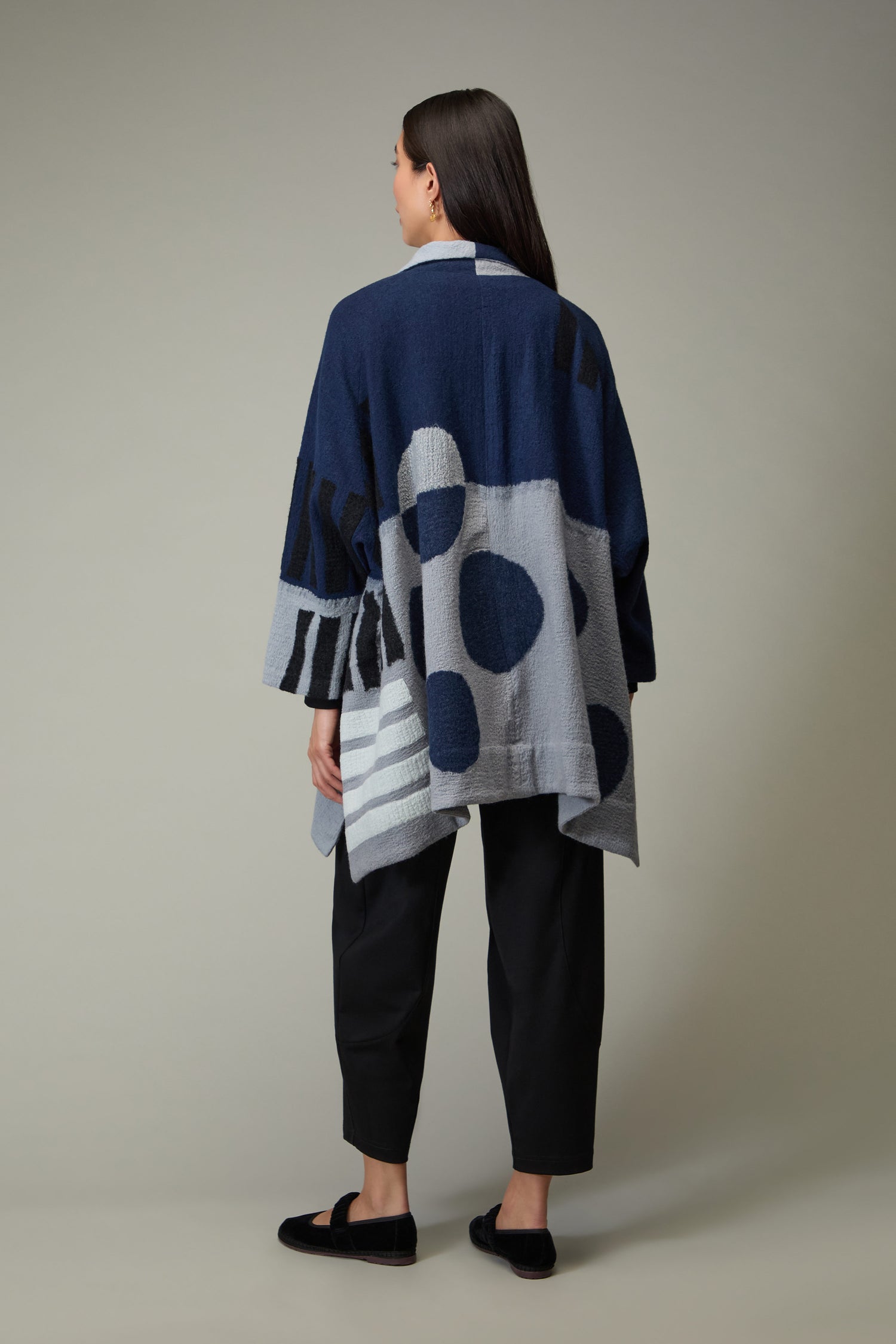 A person faces away, wearing the Spot and Dash Wool Jacket in navy and gray abstract pattern by Raga, paired with black pants and black slip-on shoes against a plain background.