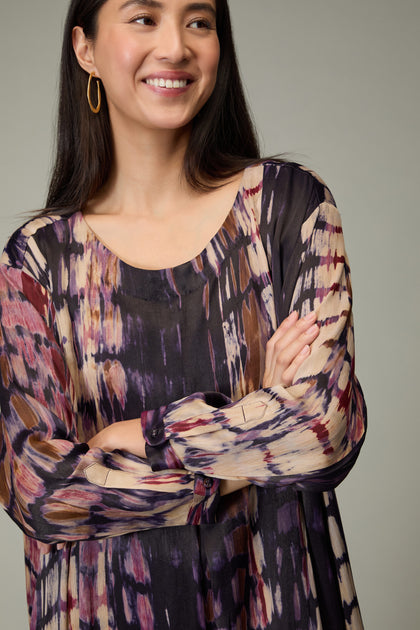 A woman with long dark hair stands with her arms crossed, smiling slightly and looking off-camera, wearing the Abstract Feather Pleat Dress.