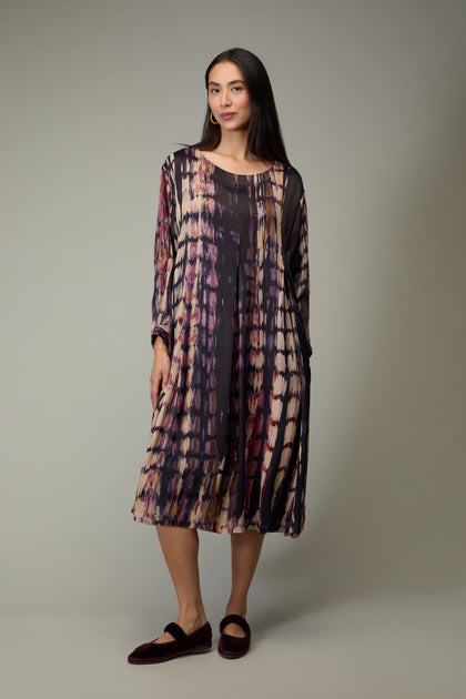 A woman wears the Abstract Feather Pleat Dress, a loose-fitting, long-sleeved piece with an abstract feather pattern in purple, burgundy, and beige hues, paired with dark slip-on shoes.