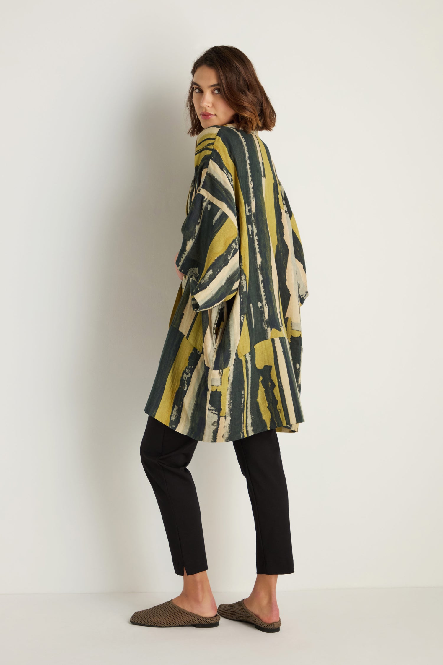 A person stands sideways against a plain white background wearing the Oversized Cotton Kimono with yellow and green painterly stripe print, paired with black pants and brown flat shoes.