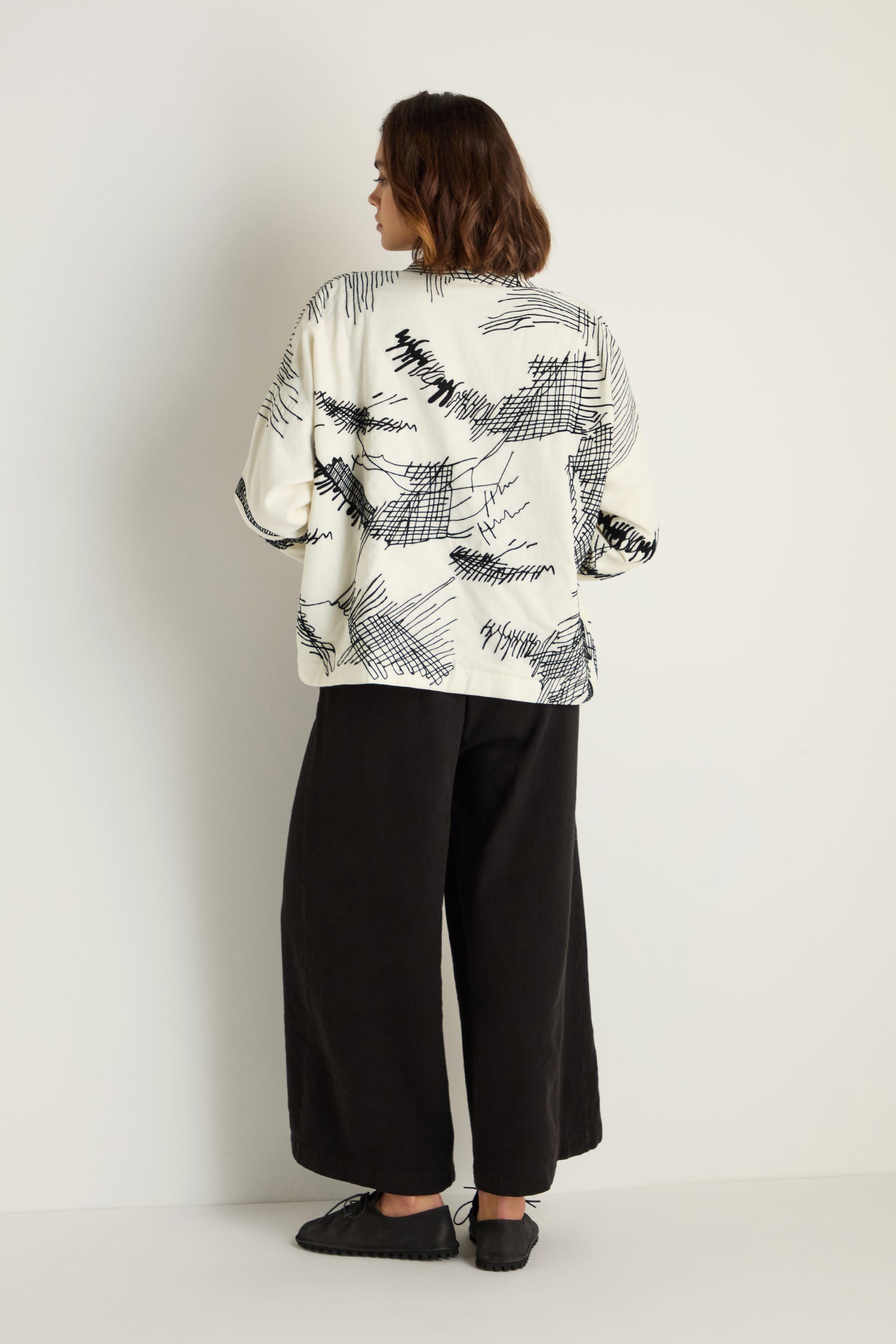 A person stands against a white wall, facing away, wearing the Embroidered Sketch Jacket with artisan black designs, paired with wide black pants and black shoes.