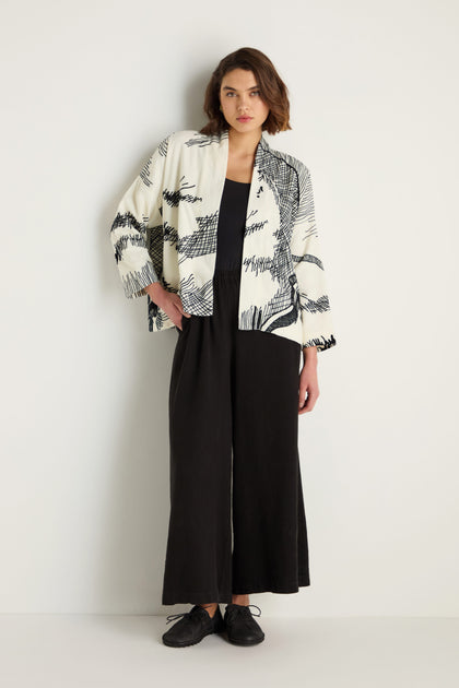 A woman stands against a plain white wall wearing a black jumpsuit, black shoes, and the Embroidered Sketch Jacket featuring white and black artisan patterns.