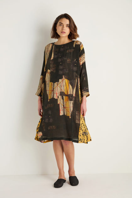 A woman stands against a white wall wearing the Klimt Print Easy Dress, featuring painterly black, brown, yellow, and beige abstract prints in a cupro viscose blend, paired with black flats.