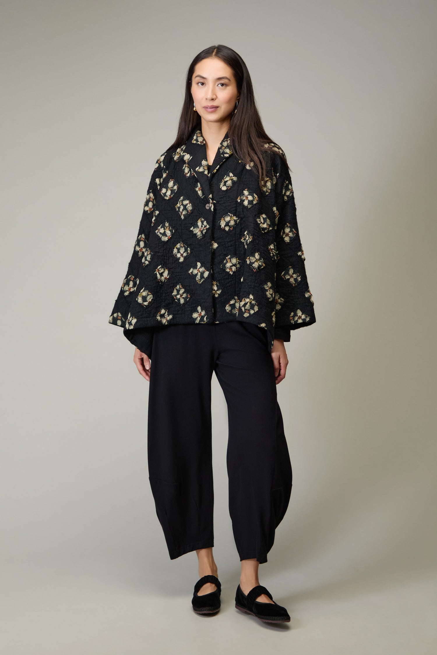 A woman stands against a plain background wearing the Textured Flower Wool Jacket, paired with loose black pants and black shoes.