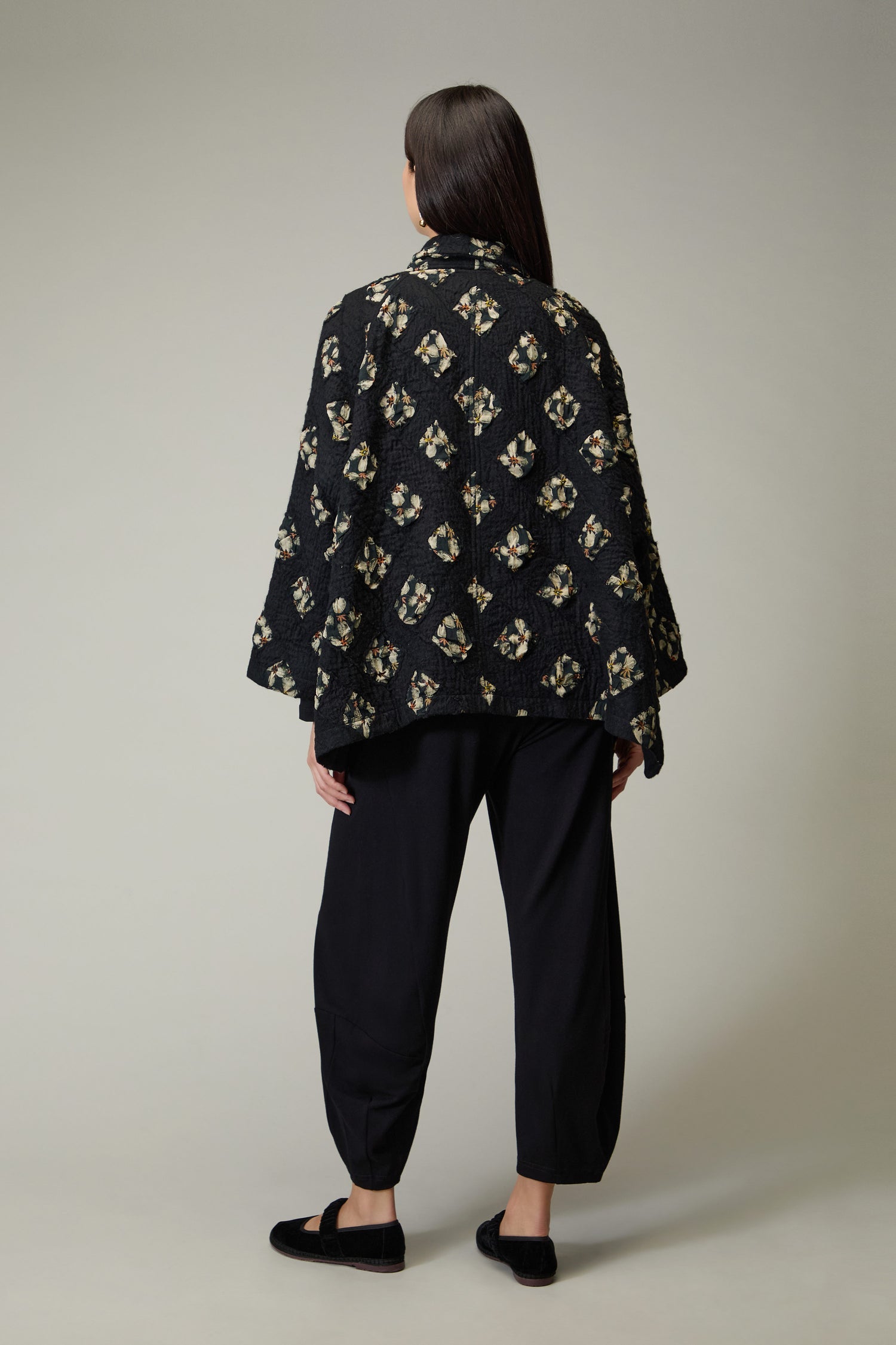 A person with long straight hair stands facing away from the camera, wearing the Textured Flower Wool Jacket, along with loose black pants and black shoes against a plain background.