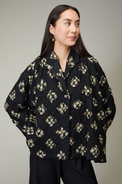 A woman stands with her hands in her pockets, wearing the Textured Flower Wool Jacket—a charcoal piece with floral designs—and black pants, looking slightly to the side.
