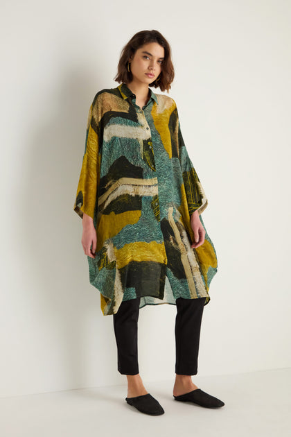 A woman stands against a plain white background wearing the Ochre Print Shirt Dress in green, yellow, and black over black pants and black shoes.