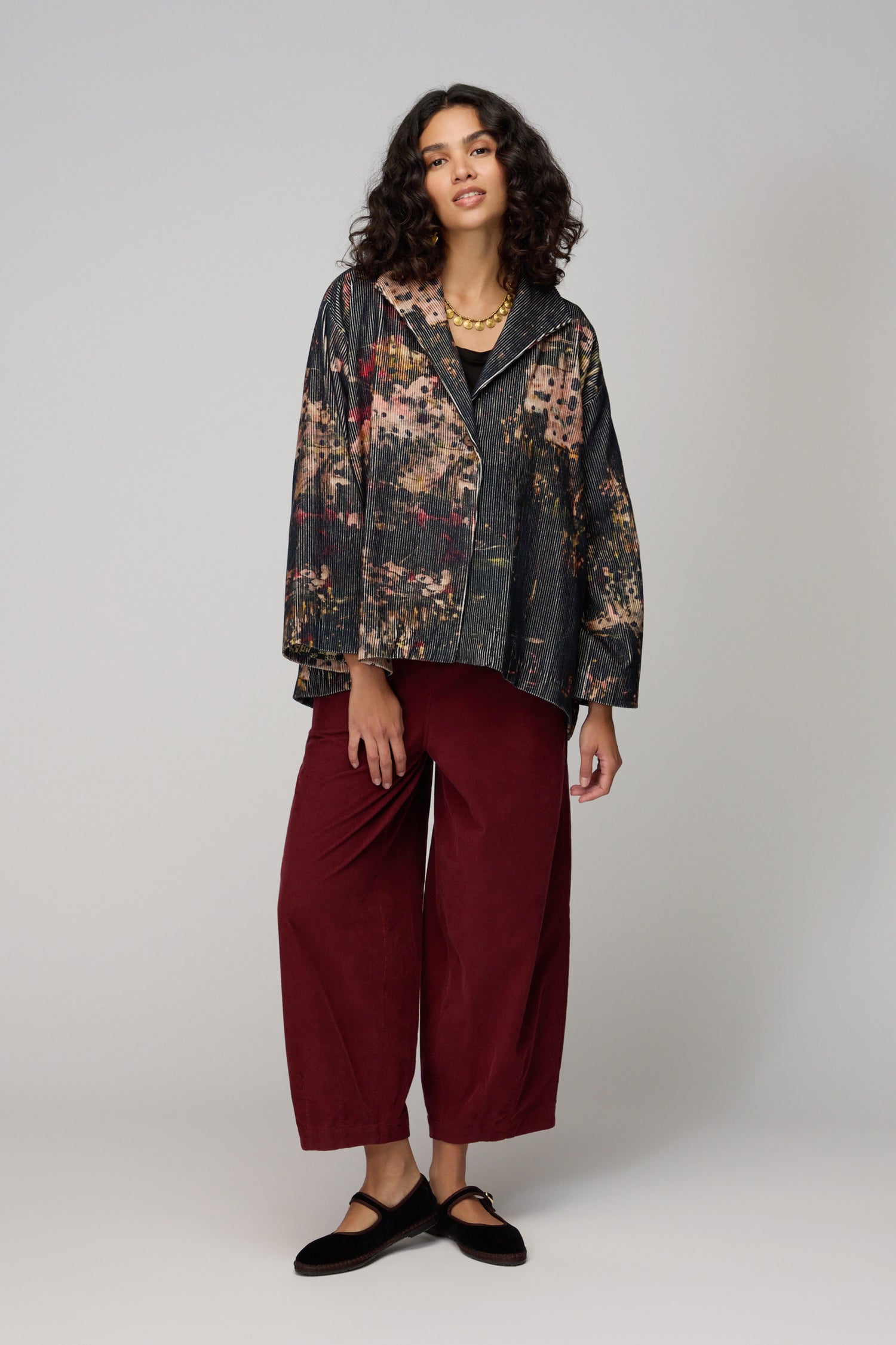 A woman stands against a plain background wearing a Print Corduroy Jacket, black top, burgundy wide-leg pants, and embellished black flats.