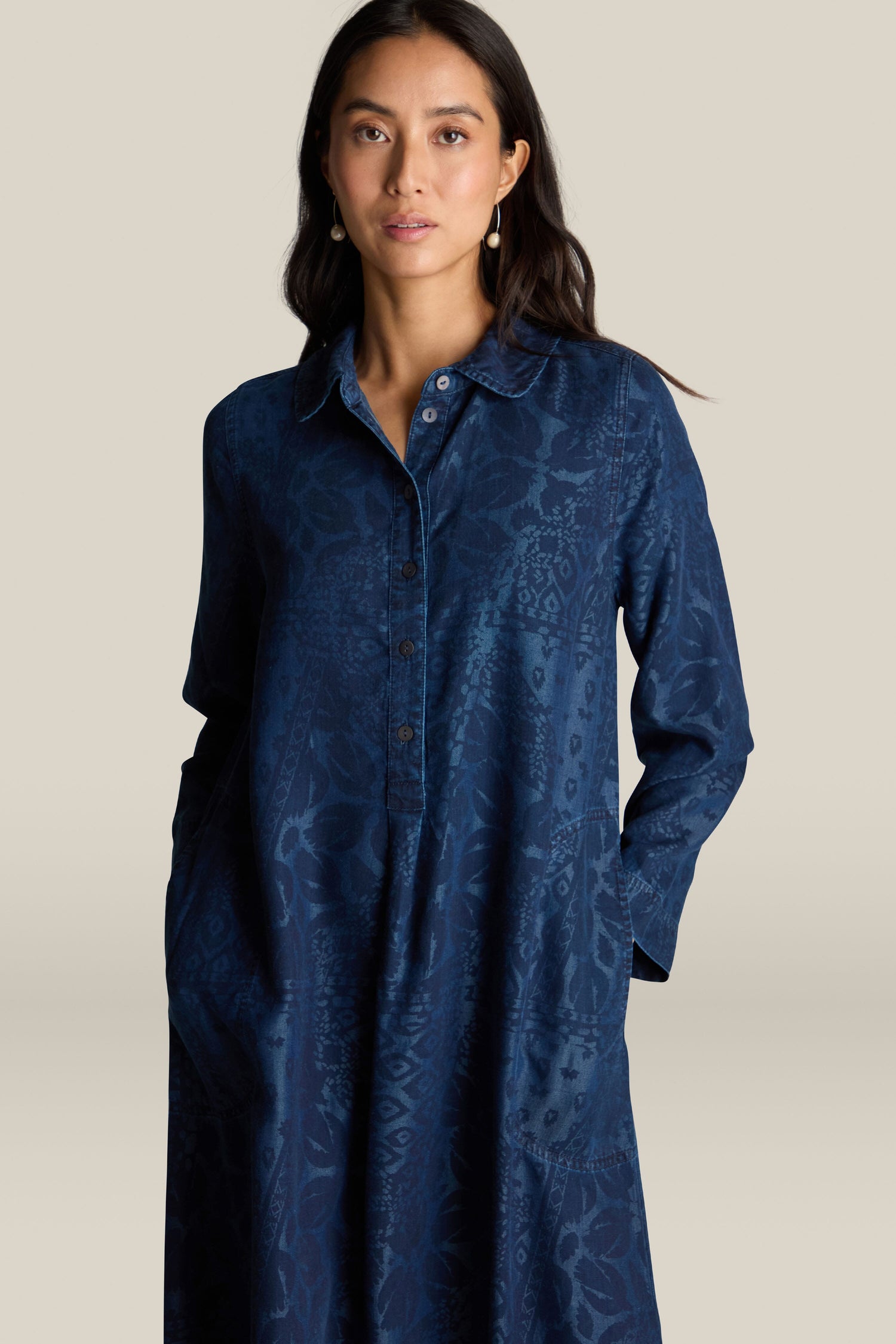 A woman stands wearing the Abstract Patchwork Print Shirt Dress, featuring long sleeves, a button-front design, and a navy blue color with a subtle abstract patchwork print. She has long dark hair and her hands are in her pockets. The background is plain and light-colored.