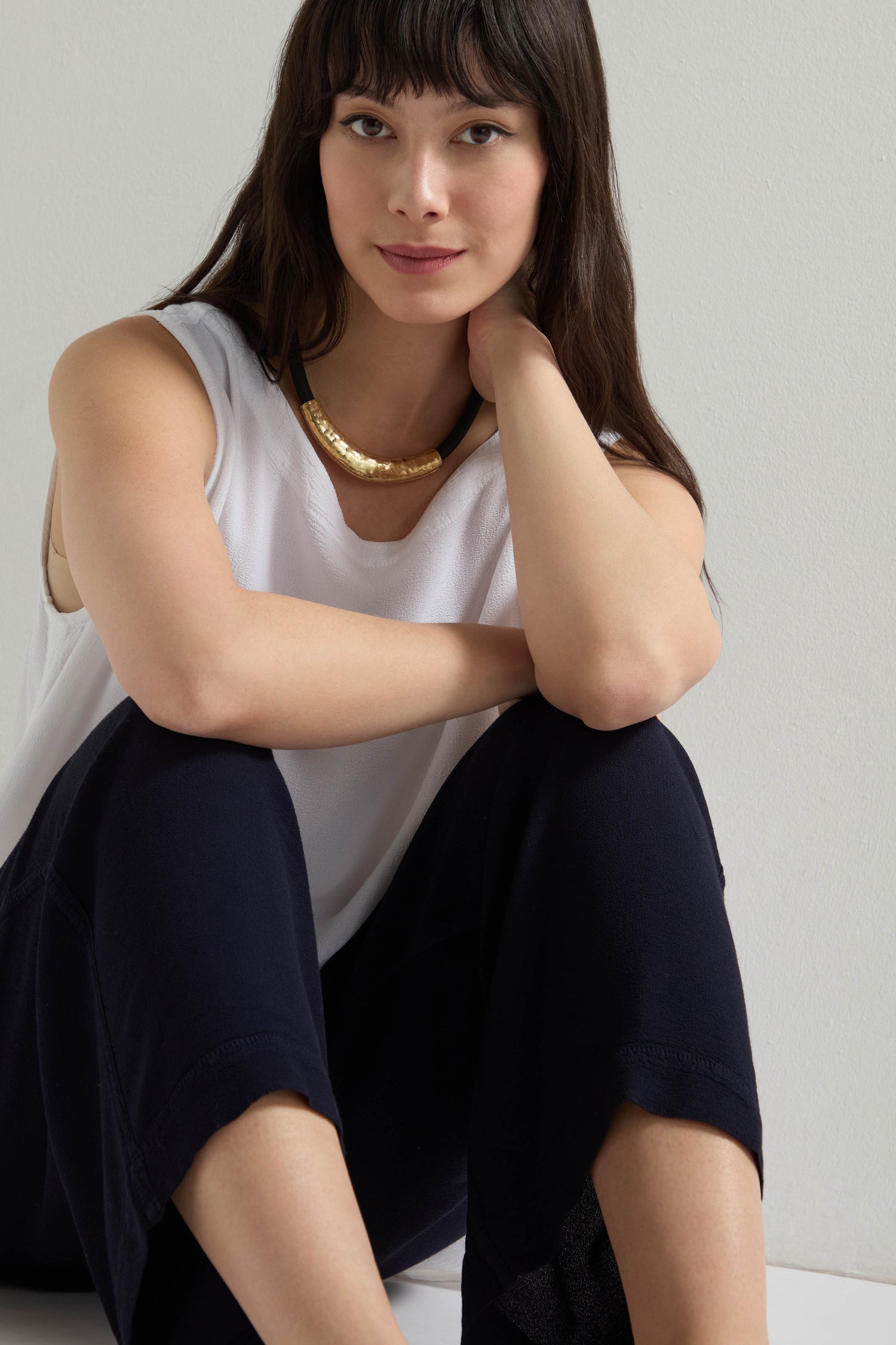 Sitting on the floor, the person wears a sleeveless white top and Crepe Wide Trousers with an elastic waistband, accented by a gold necklace. They look neutrally at the camera, their outfit embracing a chic wide-leg silhouette.