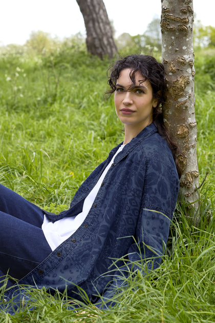 A person with curly hair sits against a tree in a grassy outdoor area, wearing an Abstract Patchwork Print Shirt beneath a blue jacket, adding a contemporary feel to the scene.