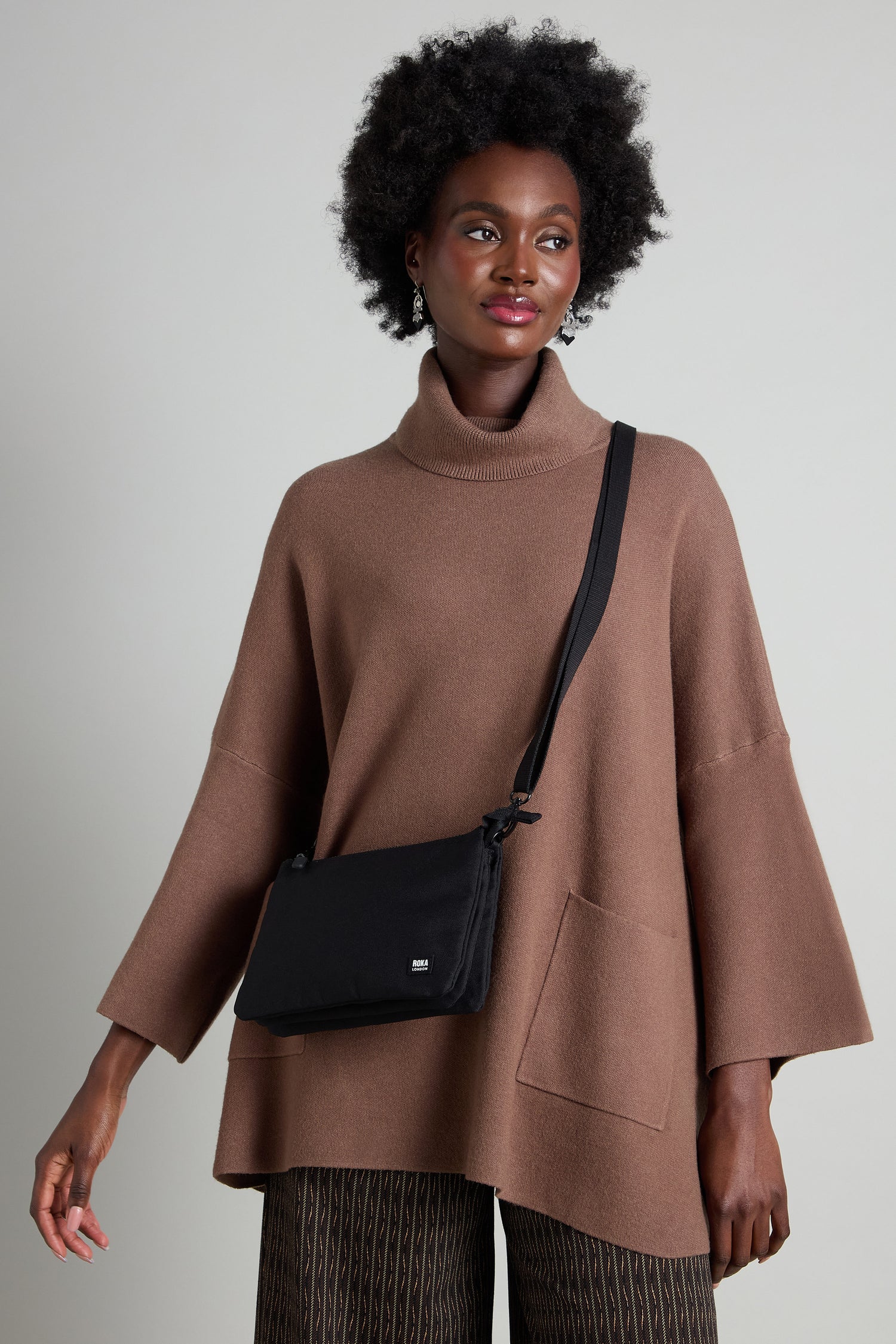 A woman in a brown turtleneck sweater, patterned pants, and the fashionable Recycled Carnaby Crossbody Bag stands against a gray background.