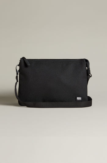 The Recycled Carnaby Crossbody Bag, made from recycled materials, features a sleek black design with a zippered top and adjustable strap, elegantly displayed against a neutral background.