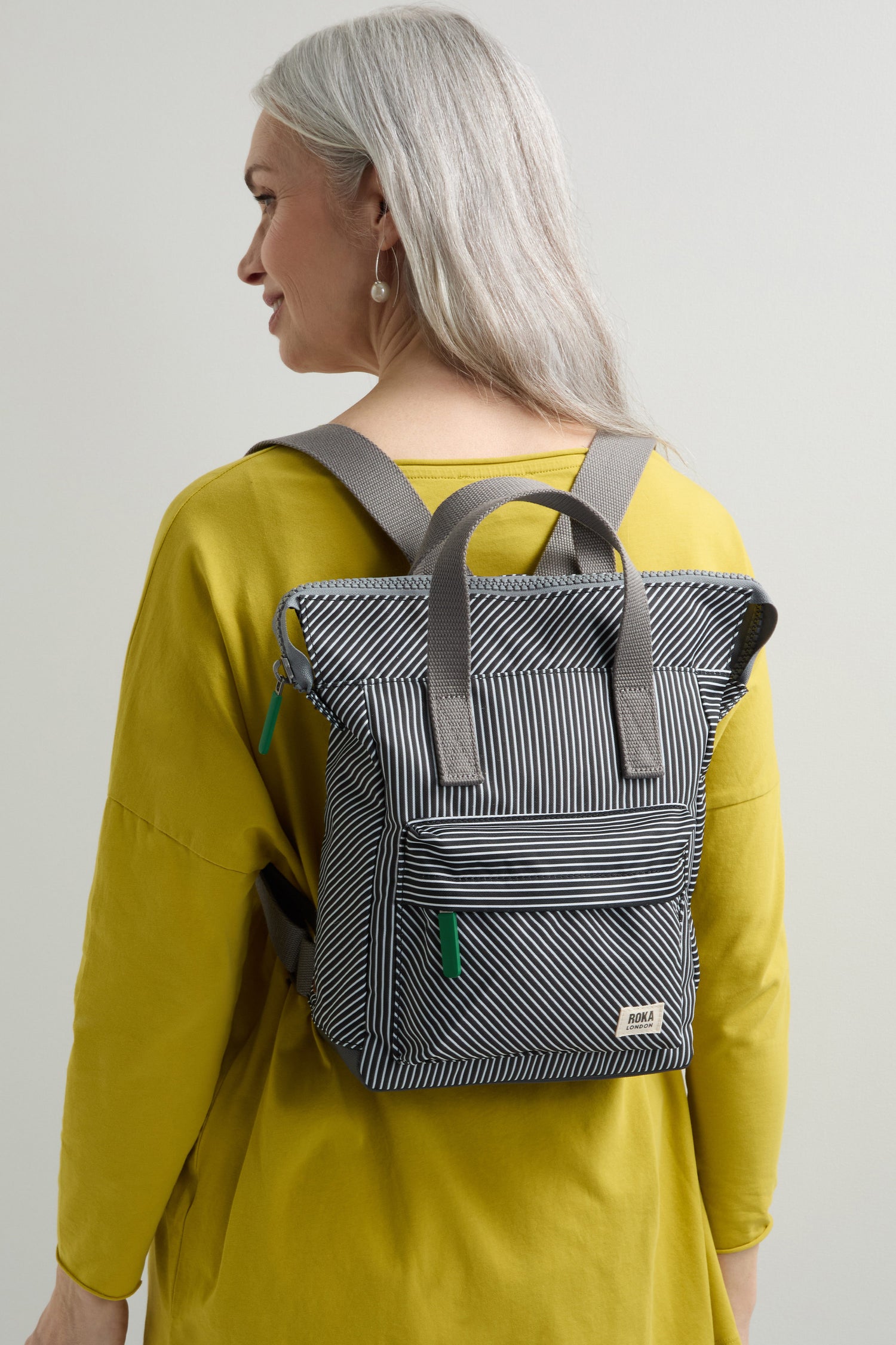 A person with long gray hair dons a yellow top, carrying the eco-conscious Recycled Canvas Stripe Bantry Bag, featuring a black-and-white striped design.