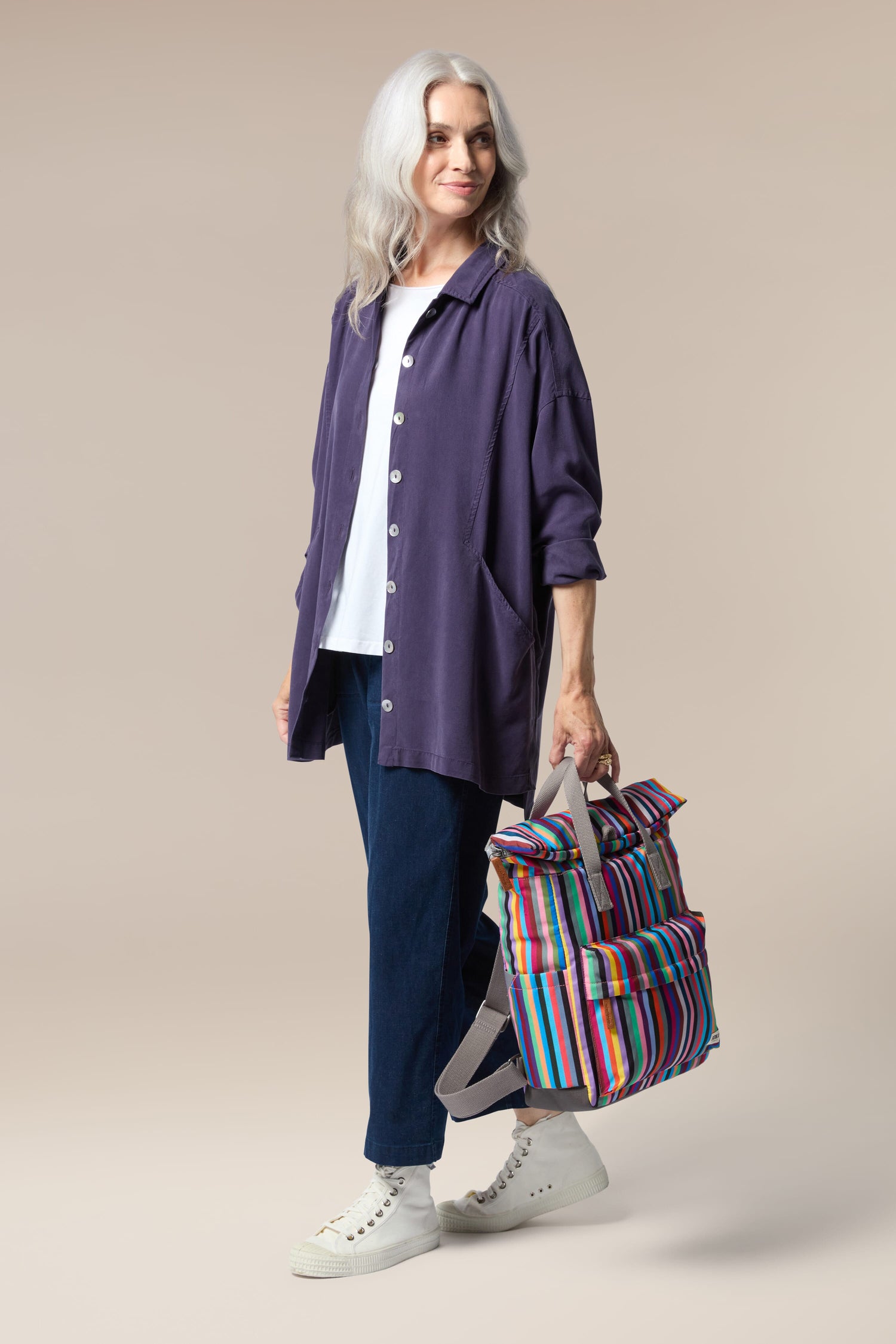 A woman wearing a purple jacket and jeans is holding a colorful Recycled Canvas Canfield Bag.