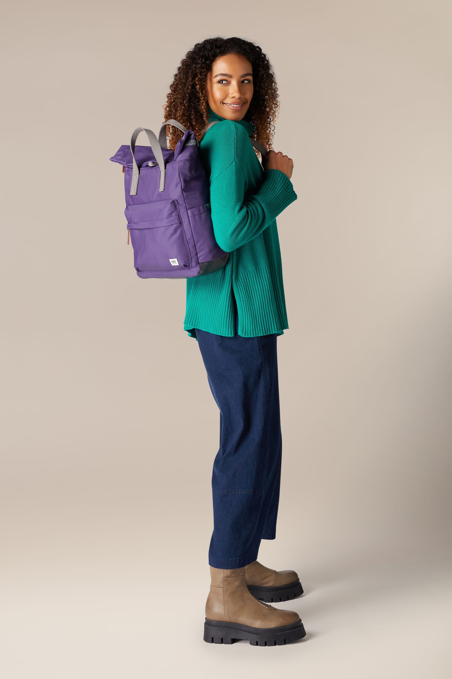 A woman wearing a purple shirt and green pants is holding a weather-resistant Recycled Canfield Bag with ample storage.