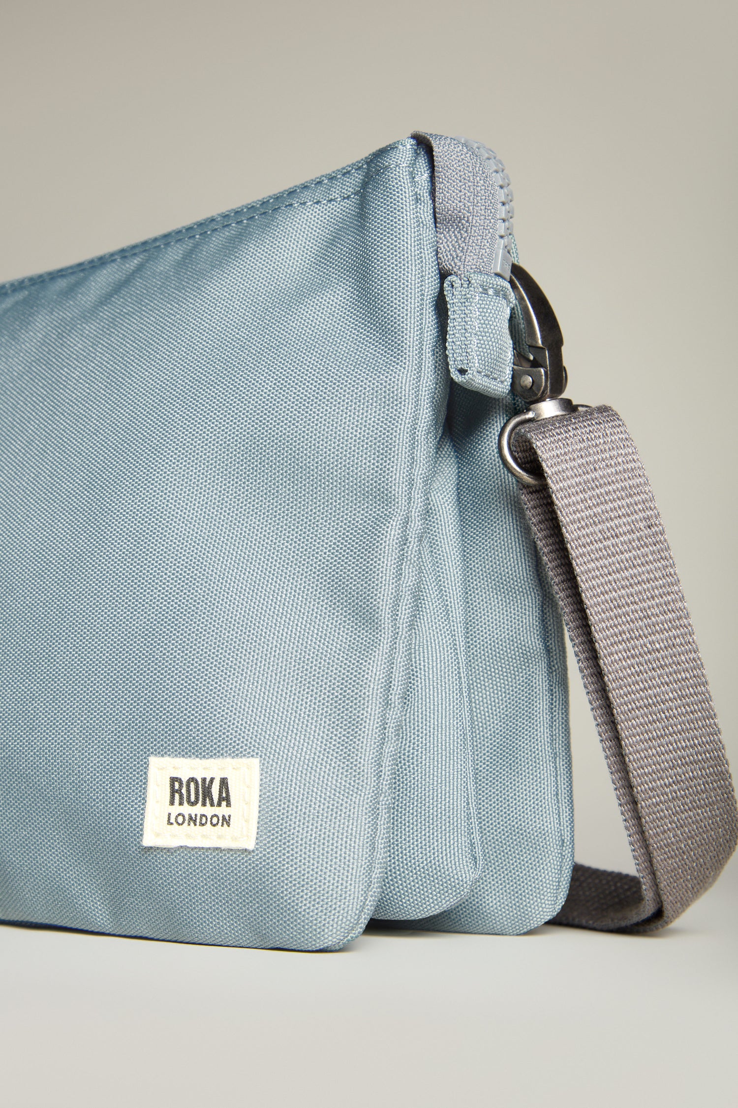 Close-up of a light blue Recycled Canvas Carnaby Crossbody Bag by Roka London, featuring a weather-resistant finish with a zipper and gray detachable strap.