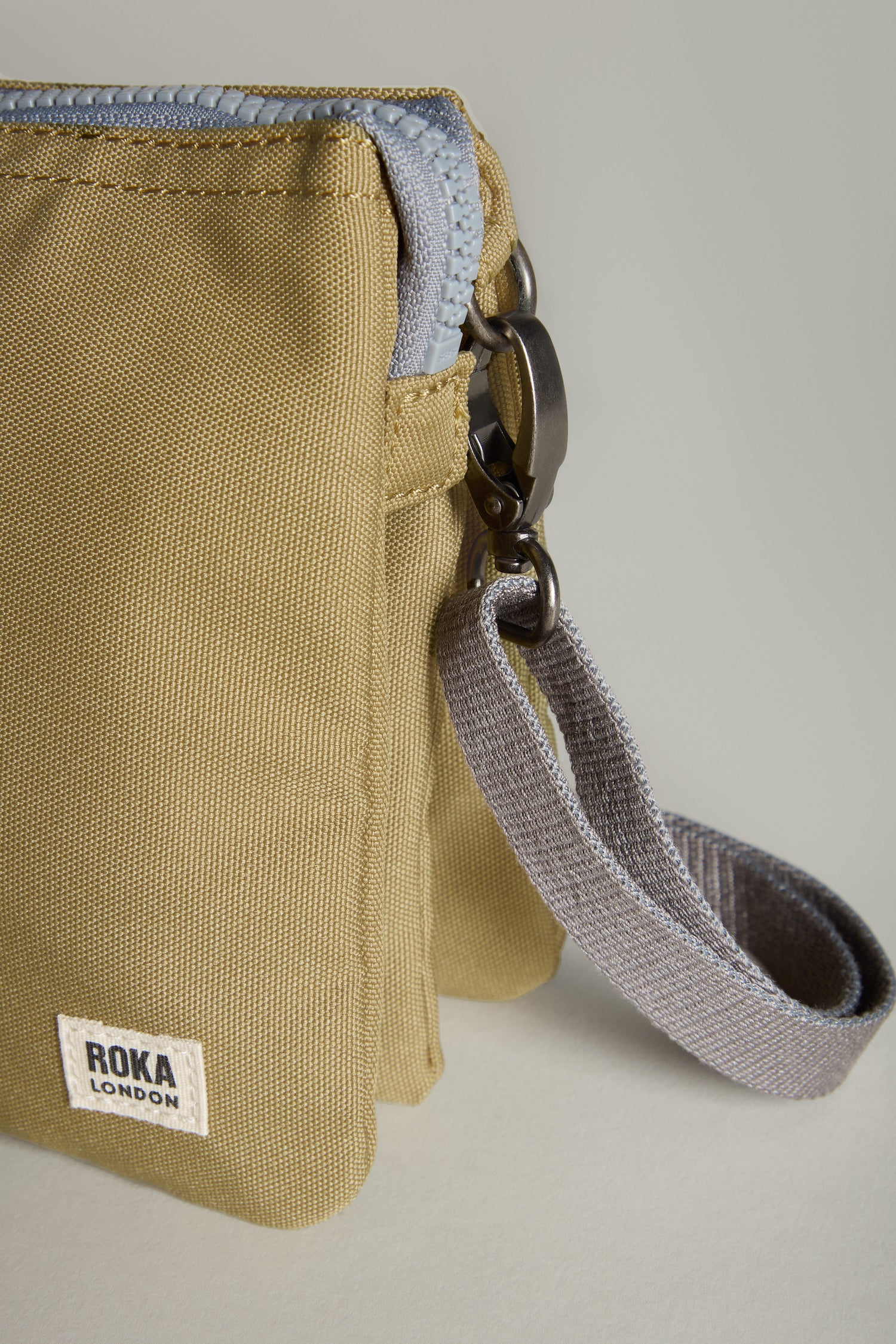 Close-up of the Recycled Canvas Carnaby Crossbody Bag from Roka London in beige, featuring a gray strap and metal clasp, set against a neutral background.