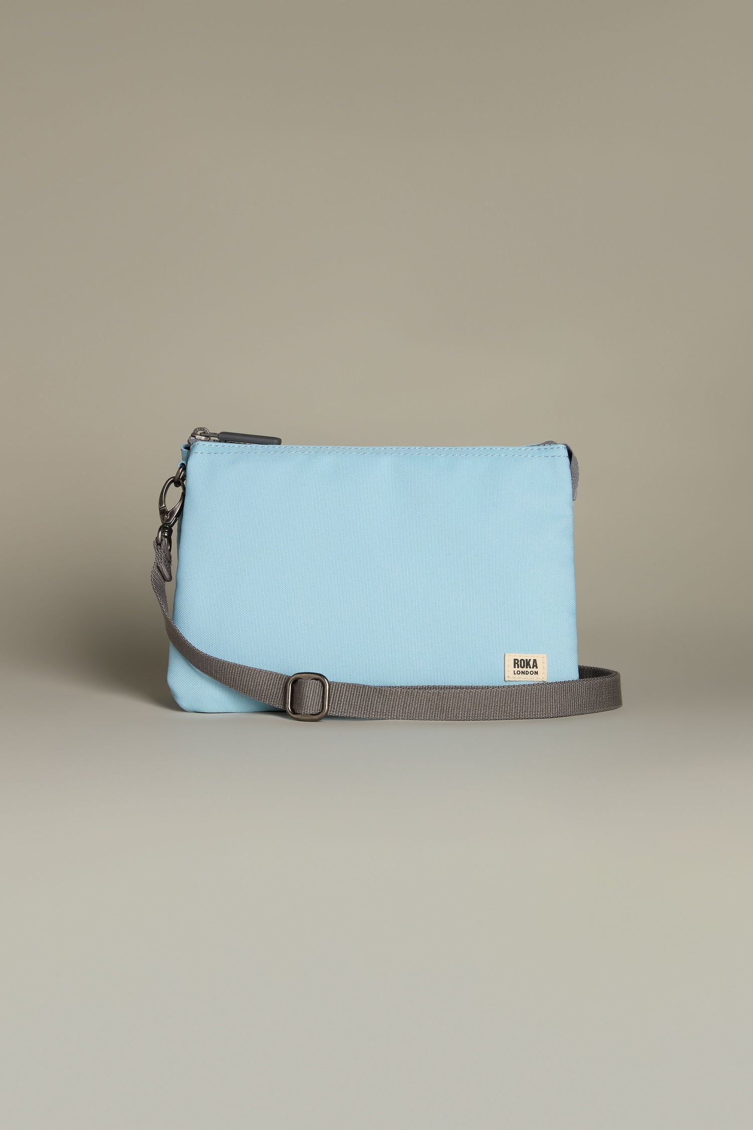 The Recycled Canvas Carnaby Crossbody Bag is light blue and rectangular, made from recycled canvas. It features a gray adjustable strap and a small logo tag on the front.