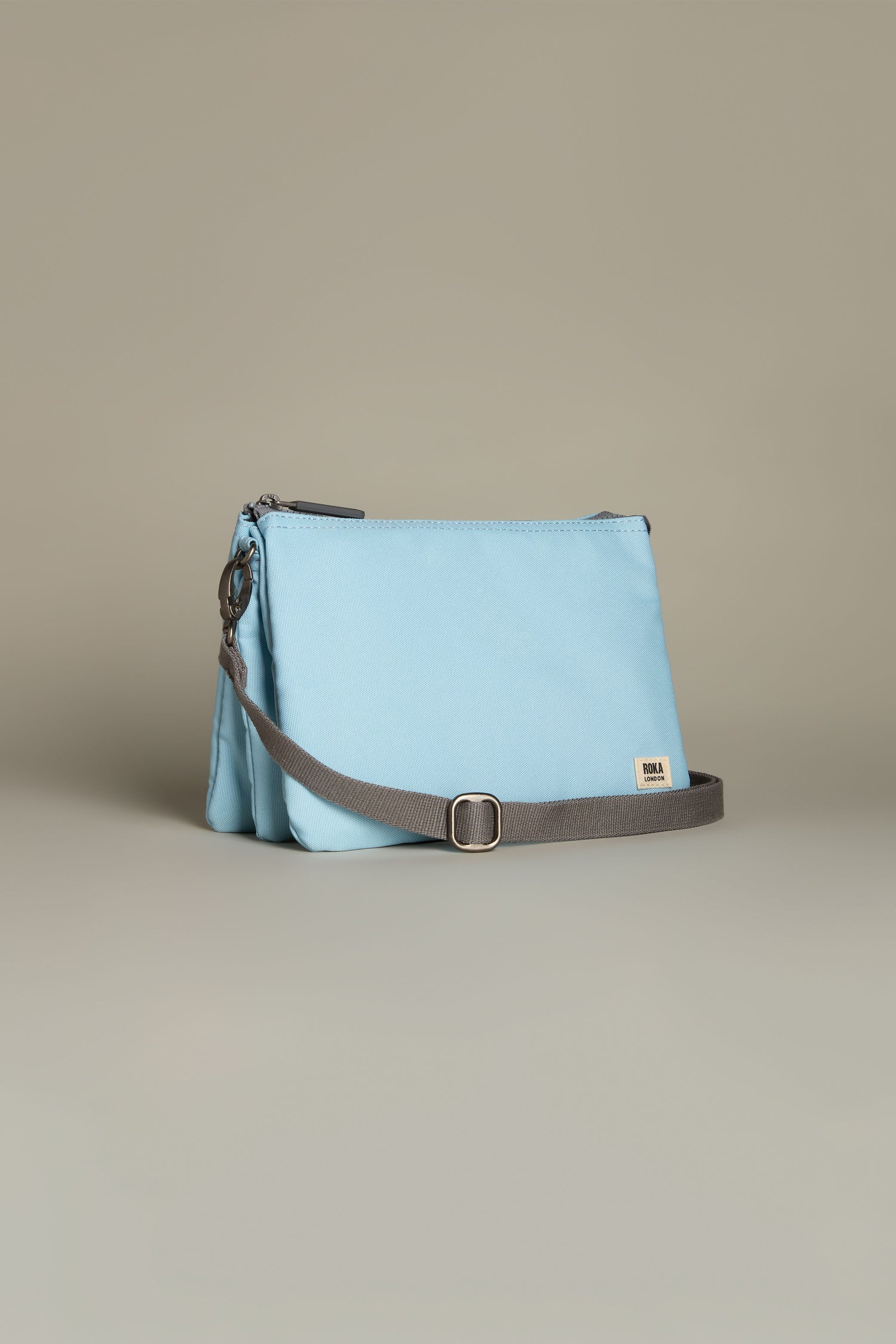 The Recycled Canvas Carnaby Crossbody Bag features a light blue design made from recycled canvas, with a gray adjustable strap and small metal buckle, set against a neutral background.