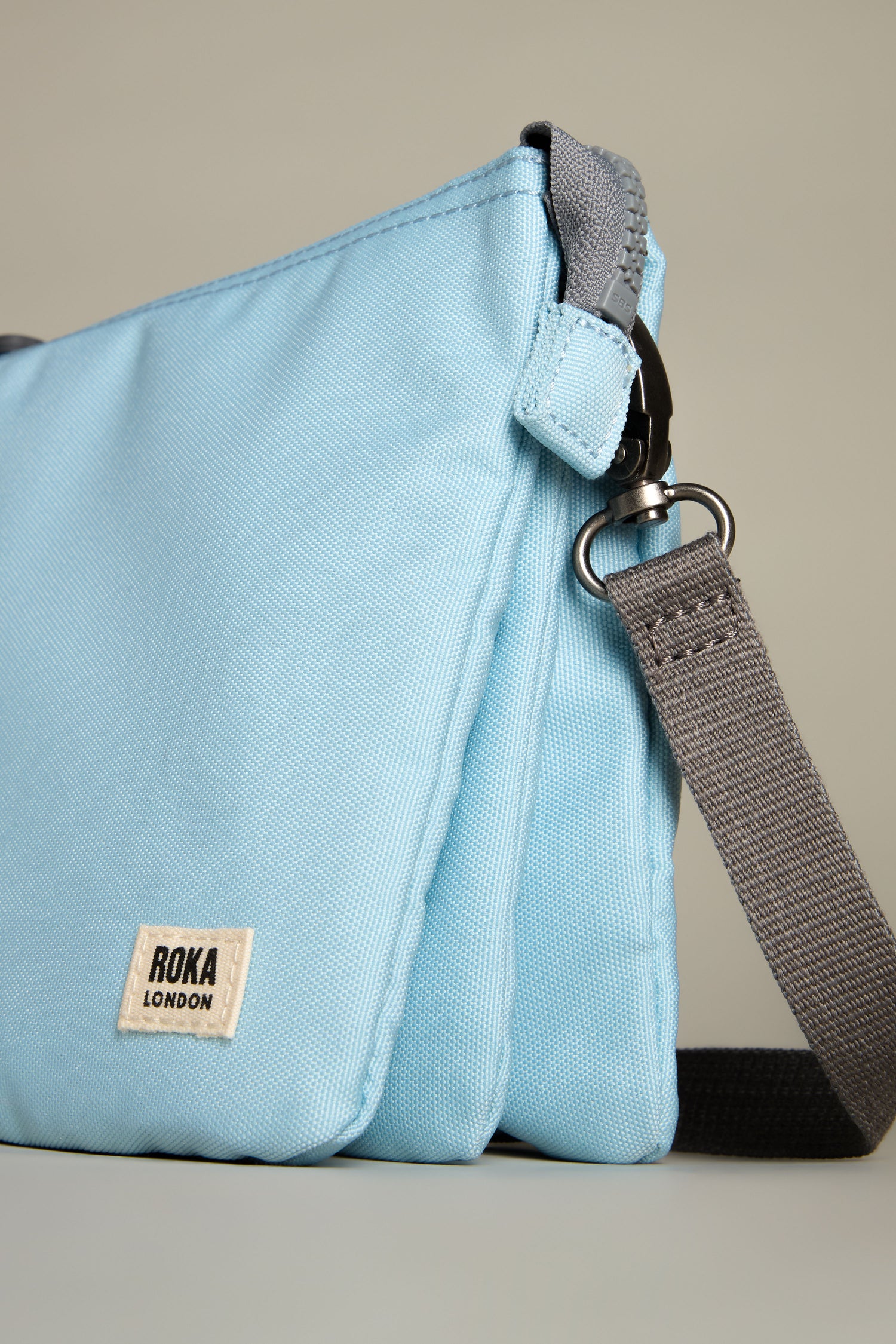 The Recycled Canvas Carnaby Crossbody Bag in light blue features a gray adjustable strap, black hardware, and a small Roka London label in the corner.