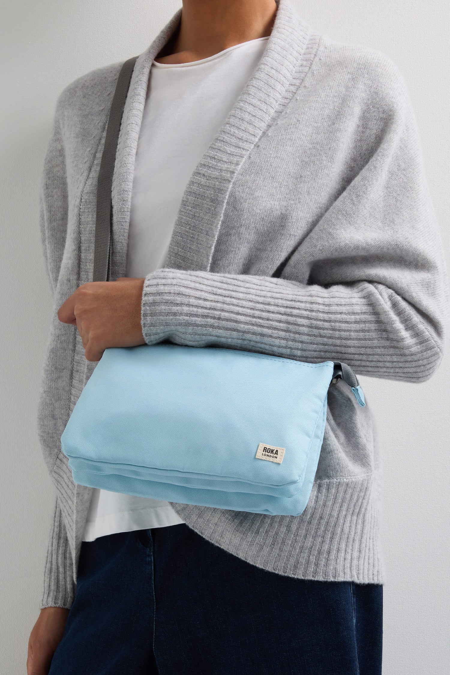 A person in a gray cardigan and white shirt holds a light blue Recycled Canvas Carnaby Crossbody, featuring an adjustable crossbody strap.