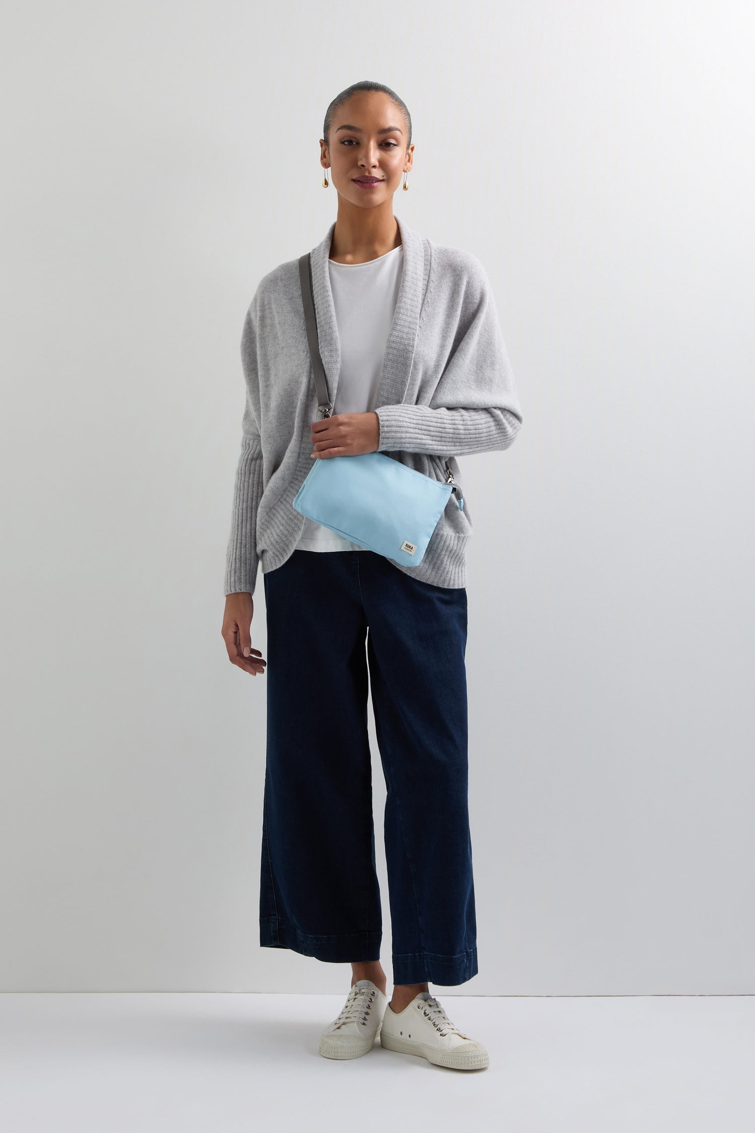 A person stands wearing a gray cardigan, white shirt, dark wide-leg pants, and white sneakers, holding a Recycled Canvas Carnaby Crossbody Bag in light blue. The bag boasts an adjustable strap and is made from recycled canvas for a chic yet sustainable look.