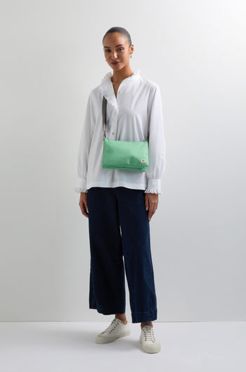 A person is against a white wall, dressed in a white blouse, dark wide-leg pants, and white sneakers. They're carrying a light green Recycled Canvas Carnaby Crossbody Bag with an adjustable strap.