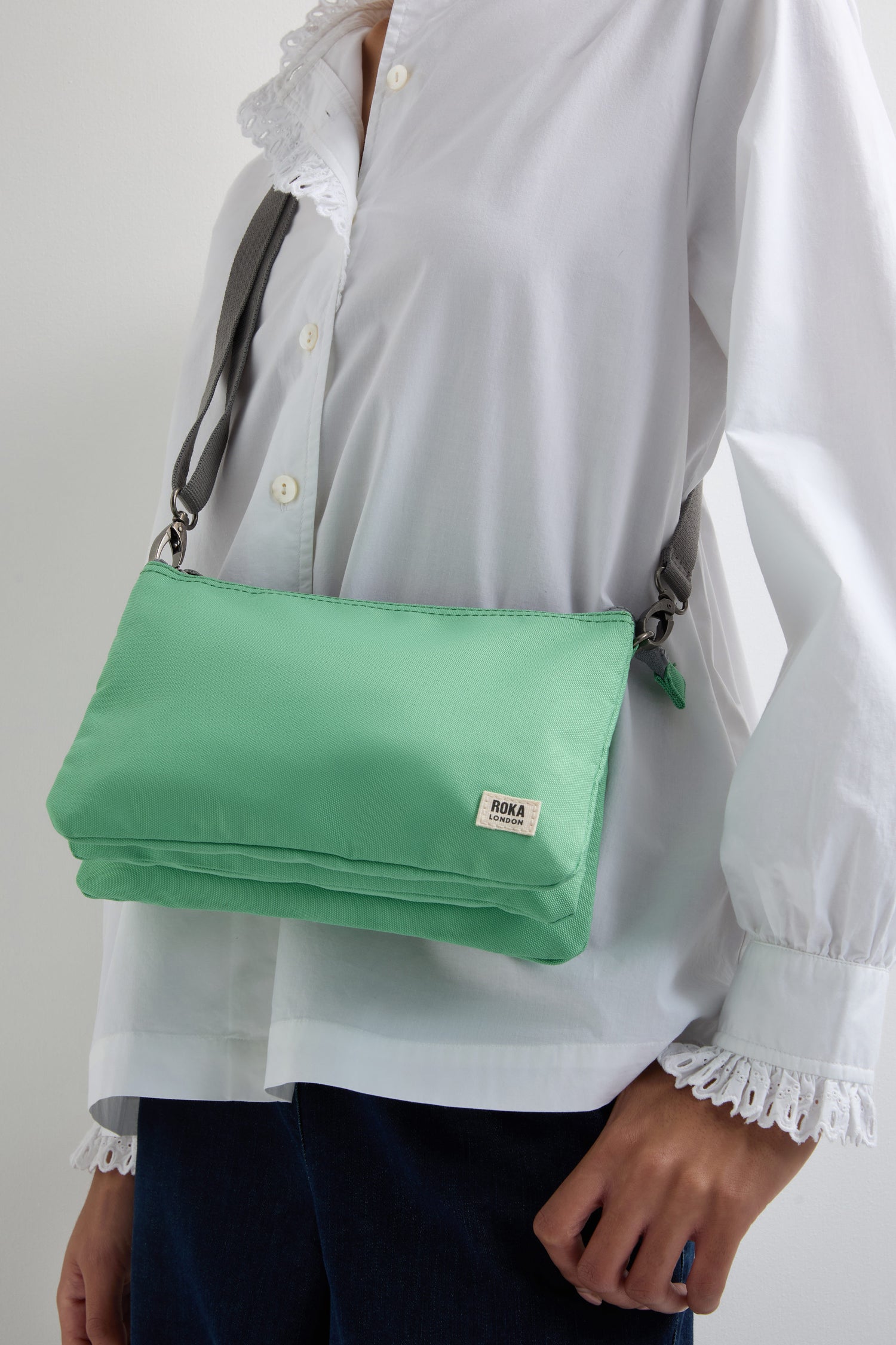 A person in a white blouse carries a Recycled Canvas Carnaby Crossbody, featuring an adjustable gray strap and chic green design.