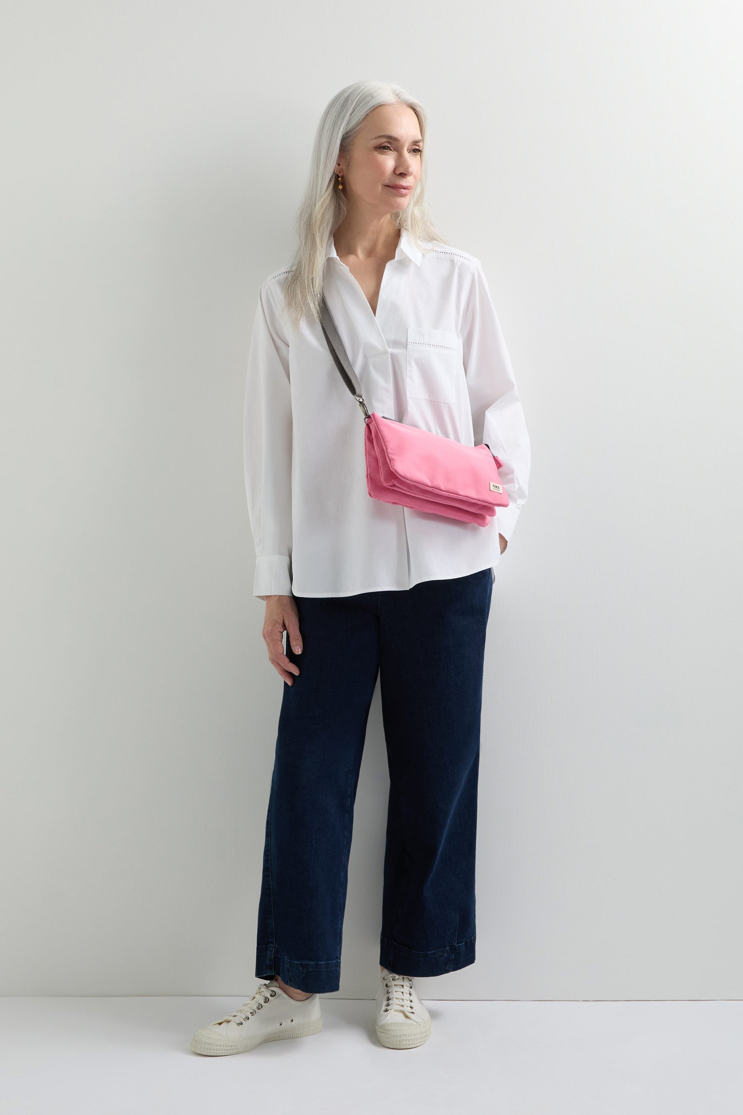 With long gray hair, wearing a white shirt and dark pants against a white background, the person sports white sneakers and carries a Recycled Canvas Carnaby Crossbody Bag with an adjustable pink crossbody strap.