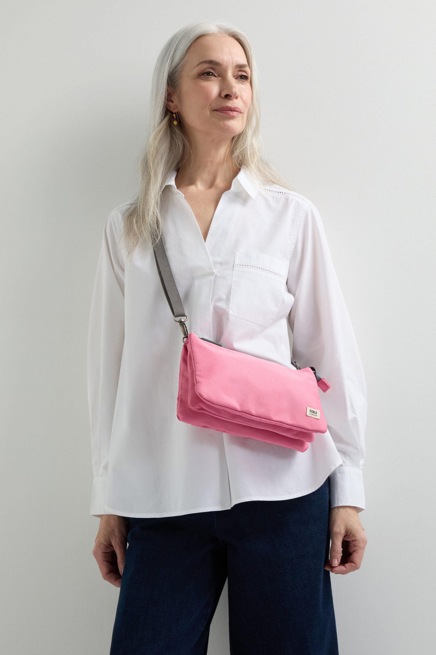 A person with long gray hair, wearing a white shirt and dark pants, casually slings the pink Recycled Canvas Carnaby Crossbody over their shoulder. The bag features an adjustable crossbody strap, perfect for versatile styling.