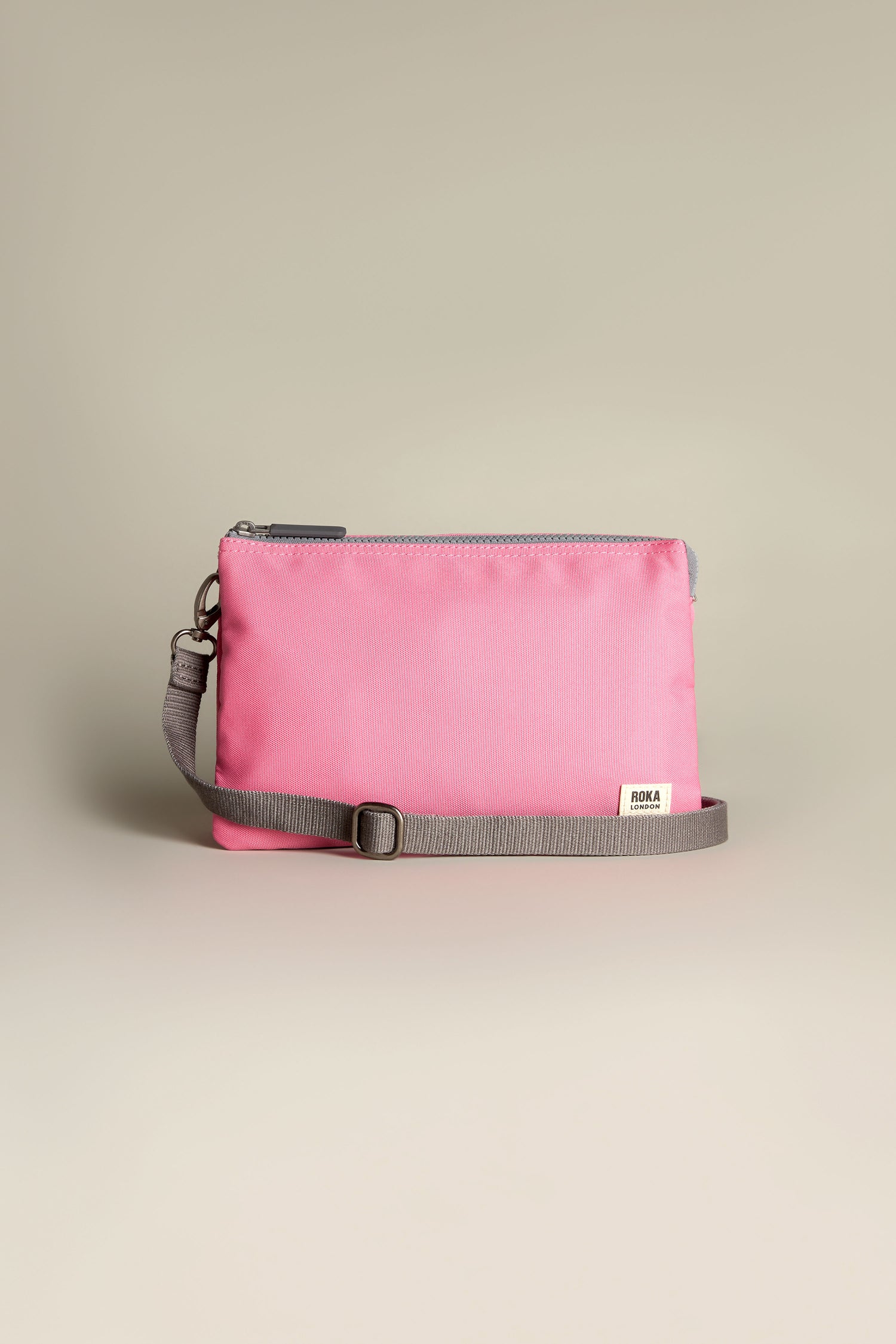 The Recycled Canvas Carnaby Crossbody Bag is a pink zipped bag resting on a neutral background, featuring a gray adjustable strap.