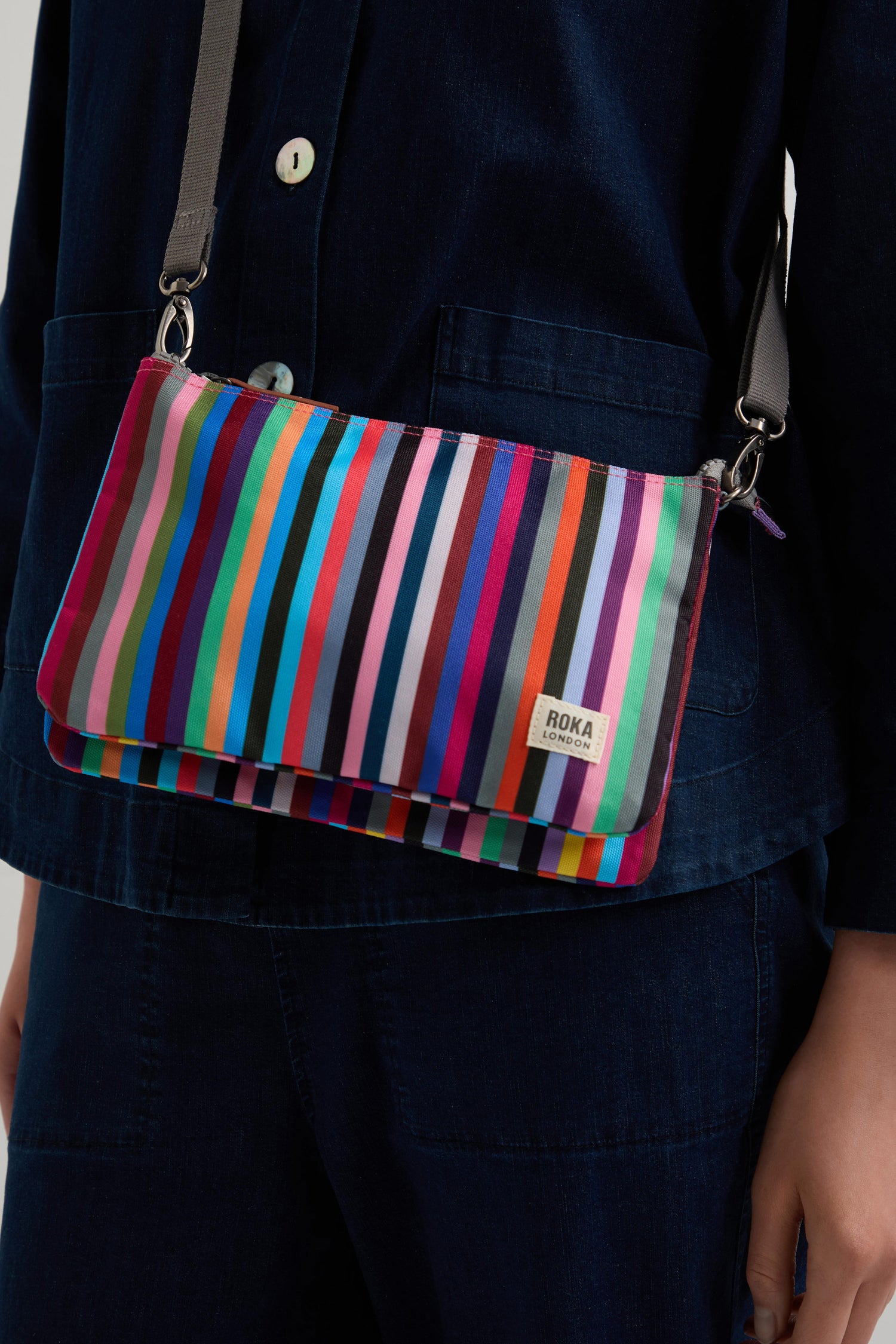 Wearing dark blue, a person showcases a bright Multi Stripe Recycled Canvas Carnaby Crossbody Bag, made from weather-resistant recycled canvas.