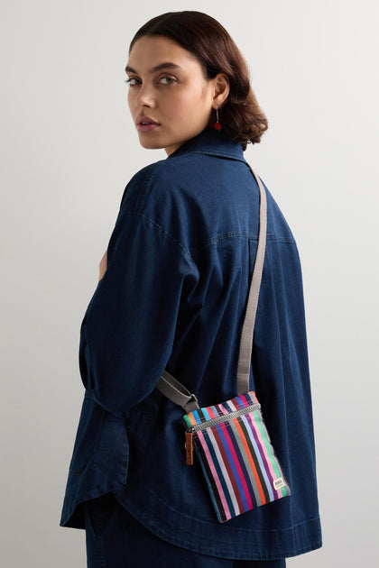 A person with short brown hair wearing a blue denim jacket holds a Recycled Chelsea Crossbody Bag, weather-resistant and striped, while looking back over their shoulder.