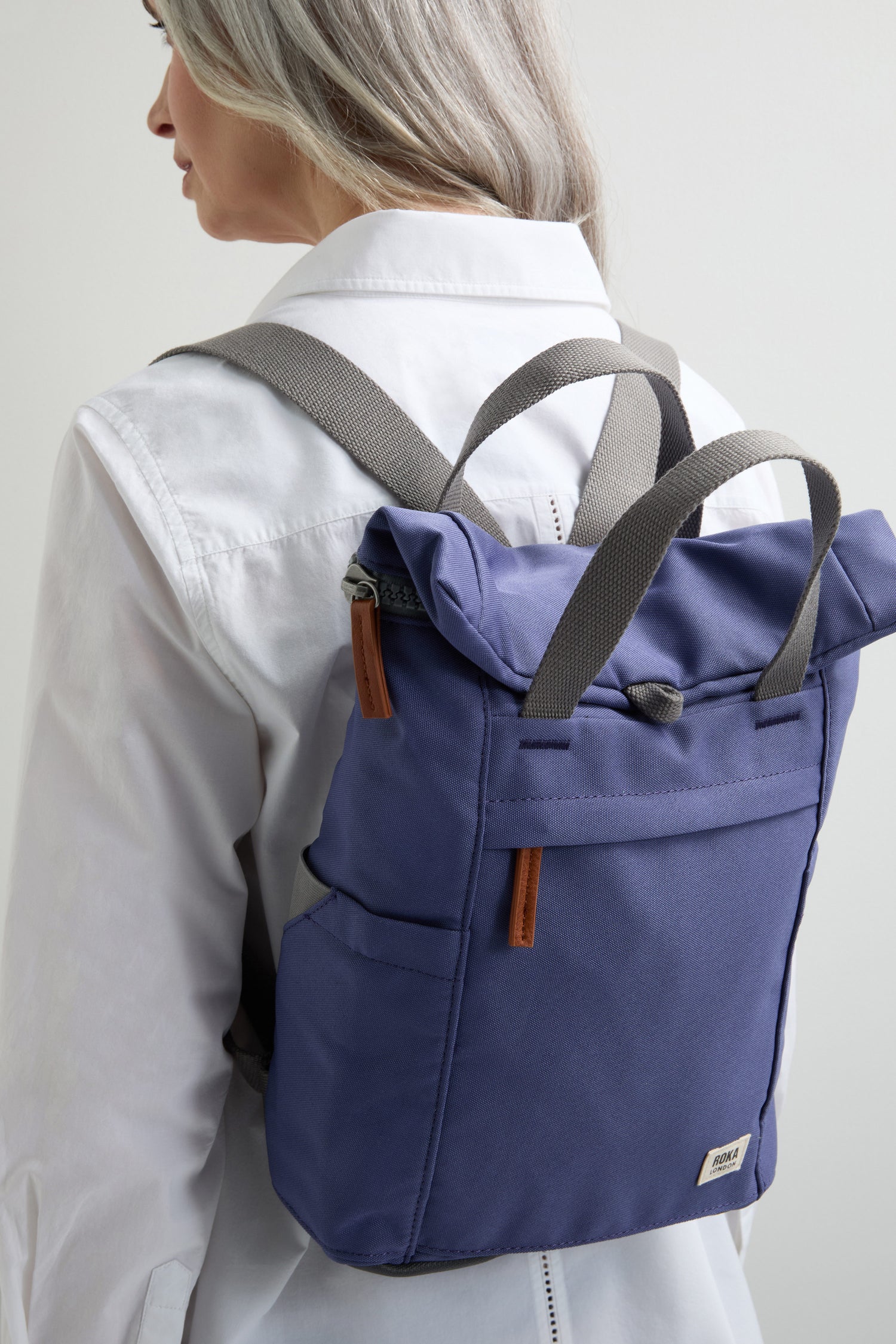 A person wears a white shirt and a purple Recycled Canvas Small Finchley Bag with gray straps, designed with eco-conscious principles. It embodies sustainable fashion while providing a stylish side view.