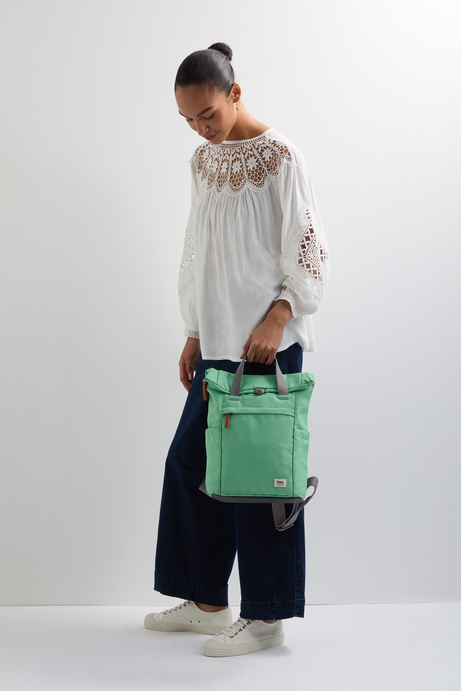 A person in a white blouse and dark pants holds the green Recycled Canvas Small Finchley Bag, embodying sustainable fashion while looking down.