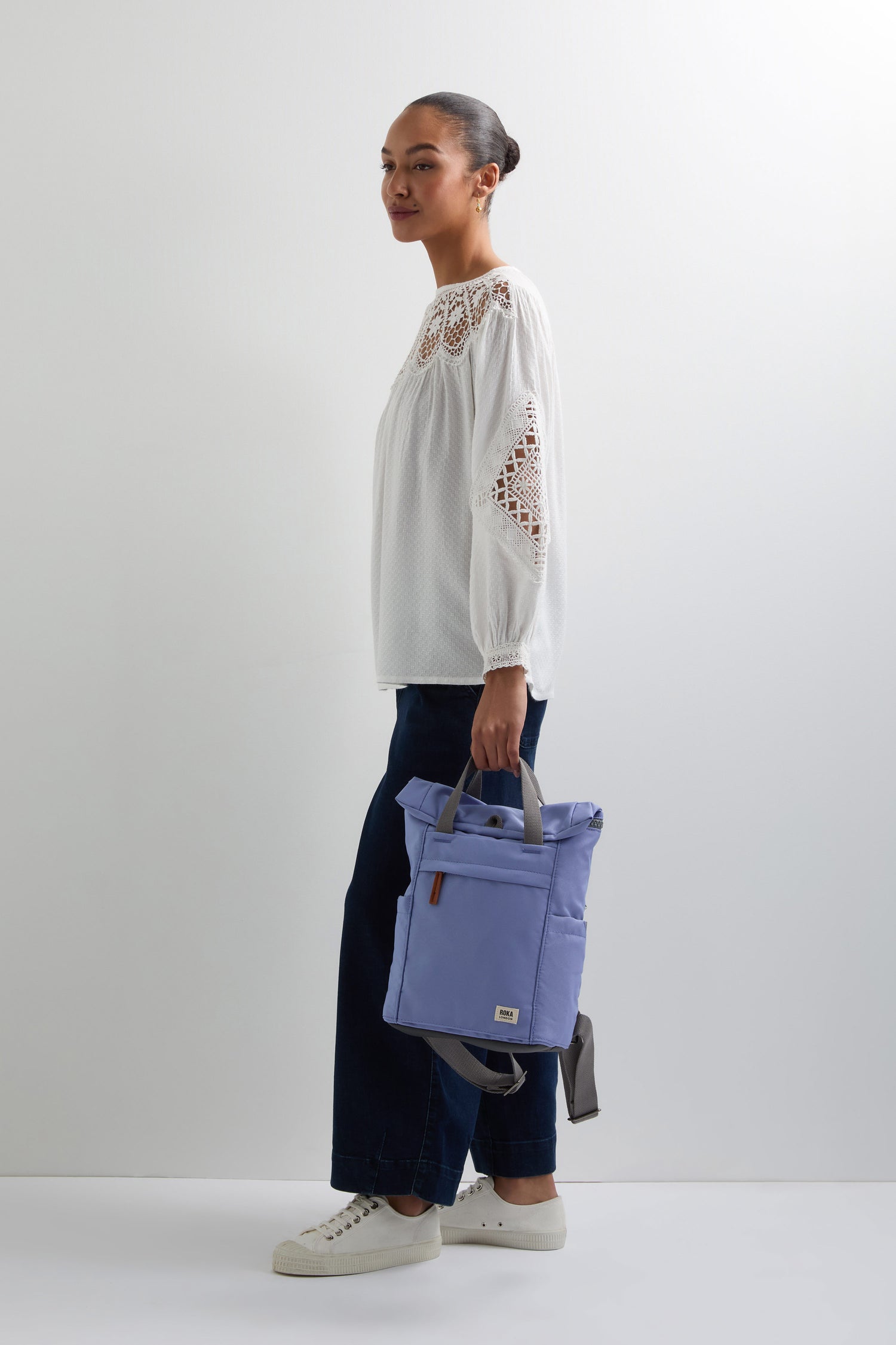 A person stands ready in a white blouse and dark pants, holding the Recycled Canvas Small Finchley Bag. This light purple bag with gray straps embodies an eco-friendly design, showcasing a dedication to sustainable fashion against a plain white backdrop.